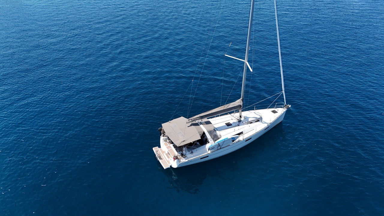Aerial view looking down on a Beneteau Oceanis 45 sailing yacht anchored in deep blue water, showing its wide cockpit bimini, swim platform, and wind generator mounted on the stern arch.