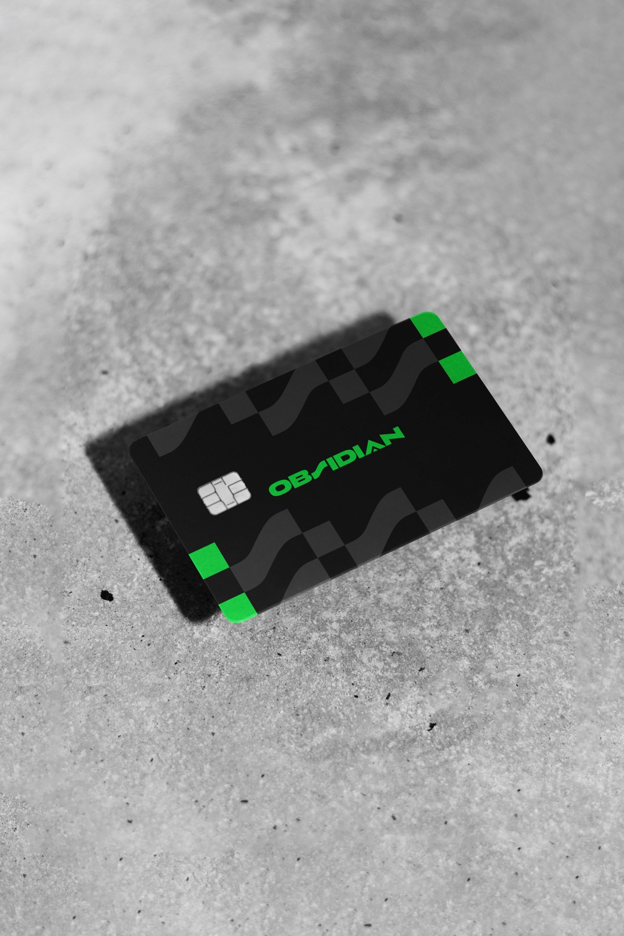 A black rectangular card with green bands along its edges lies on a gray concrete surface. The card displays the word "OBIDIAN" in green text near the top center. A small white chip is visible near the top left corner. The surface shows texture and minor imperfections.