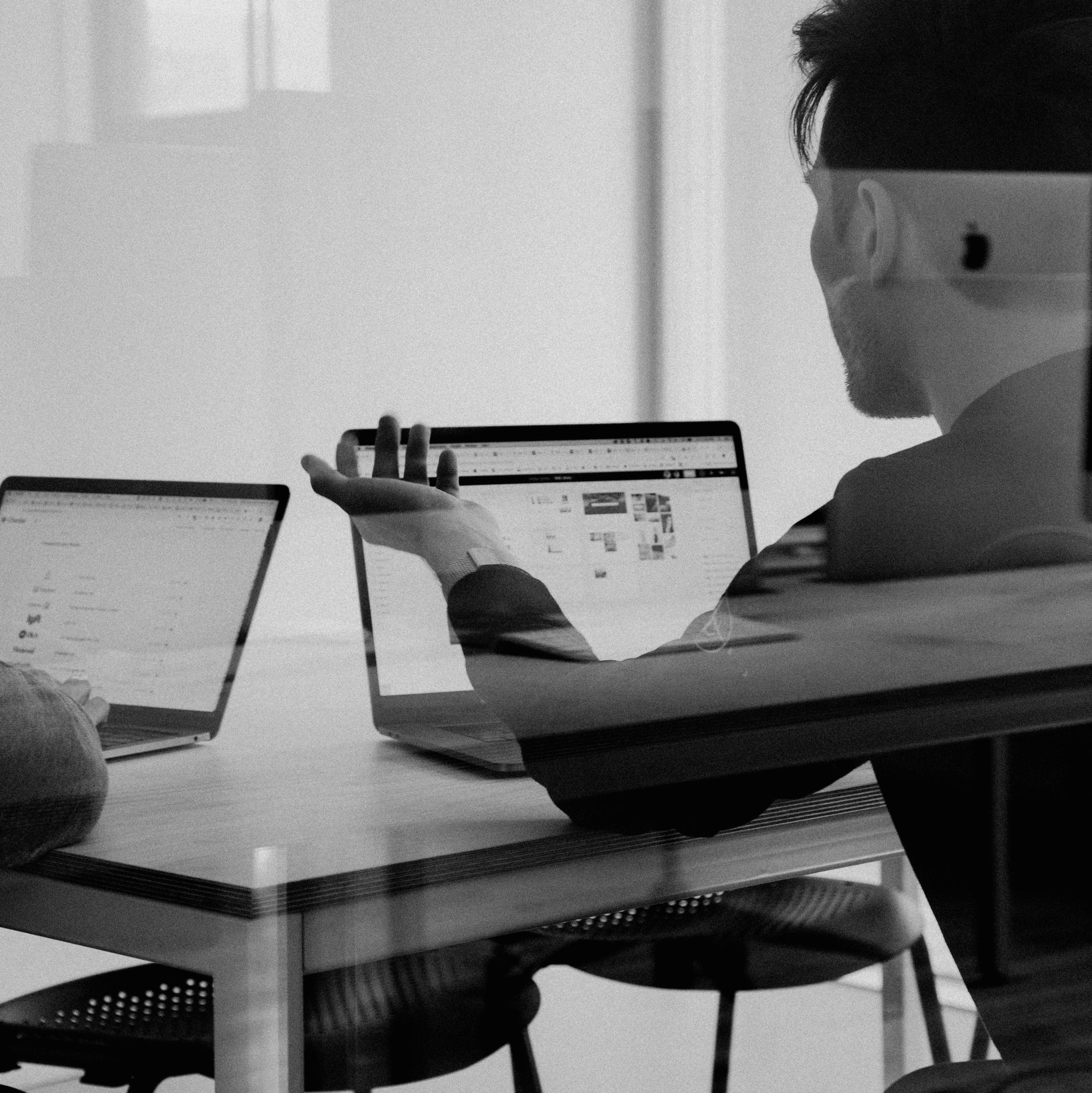 A young person with curly hair smiles at the camera while sitting in front of a laptop against a dark background.