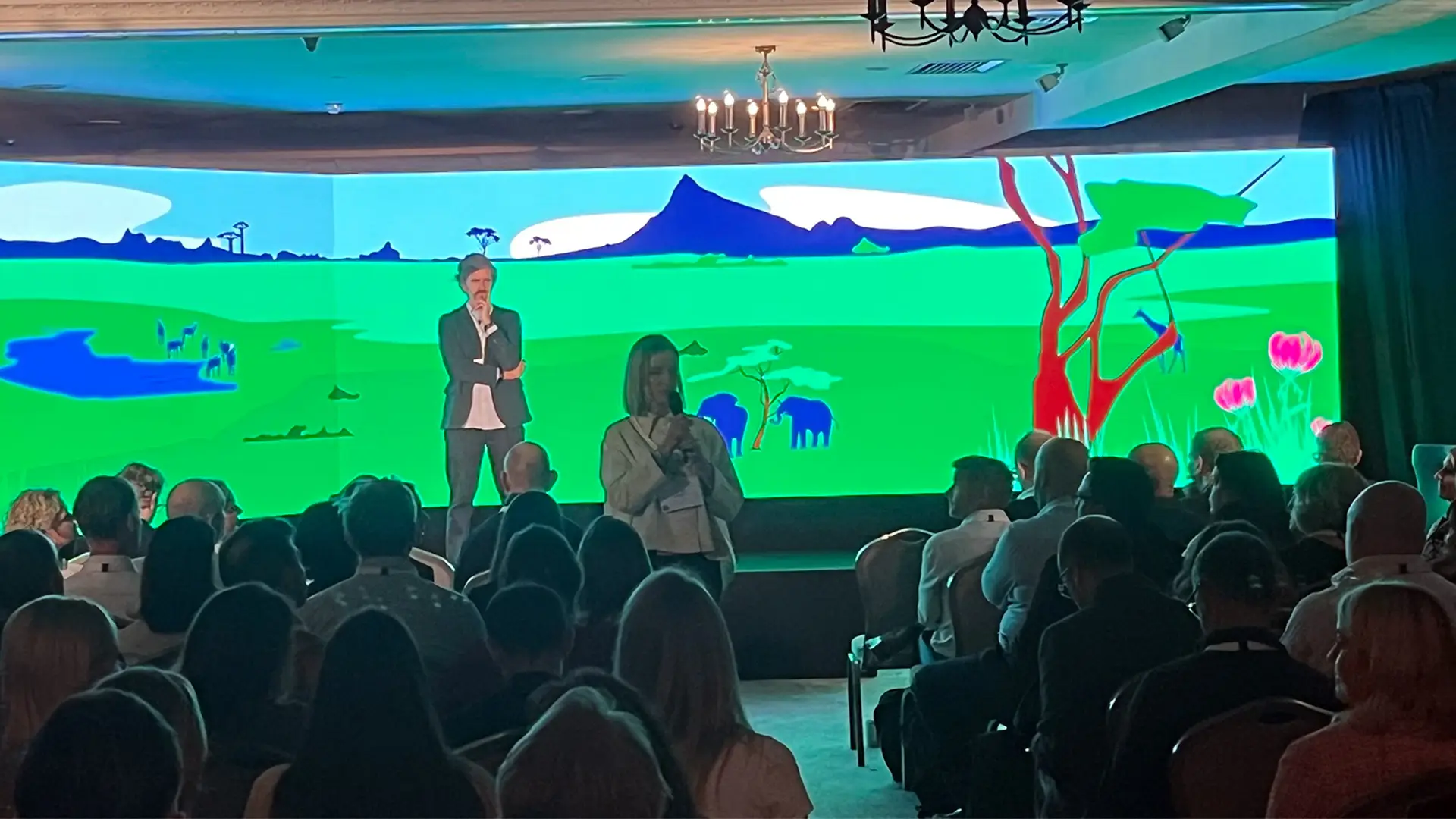 Audience member speaking into a microphone during a Bupa Strategy Cascades event, with a large illustrated landscape backdrop on stage and delegates seated in the audience.
