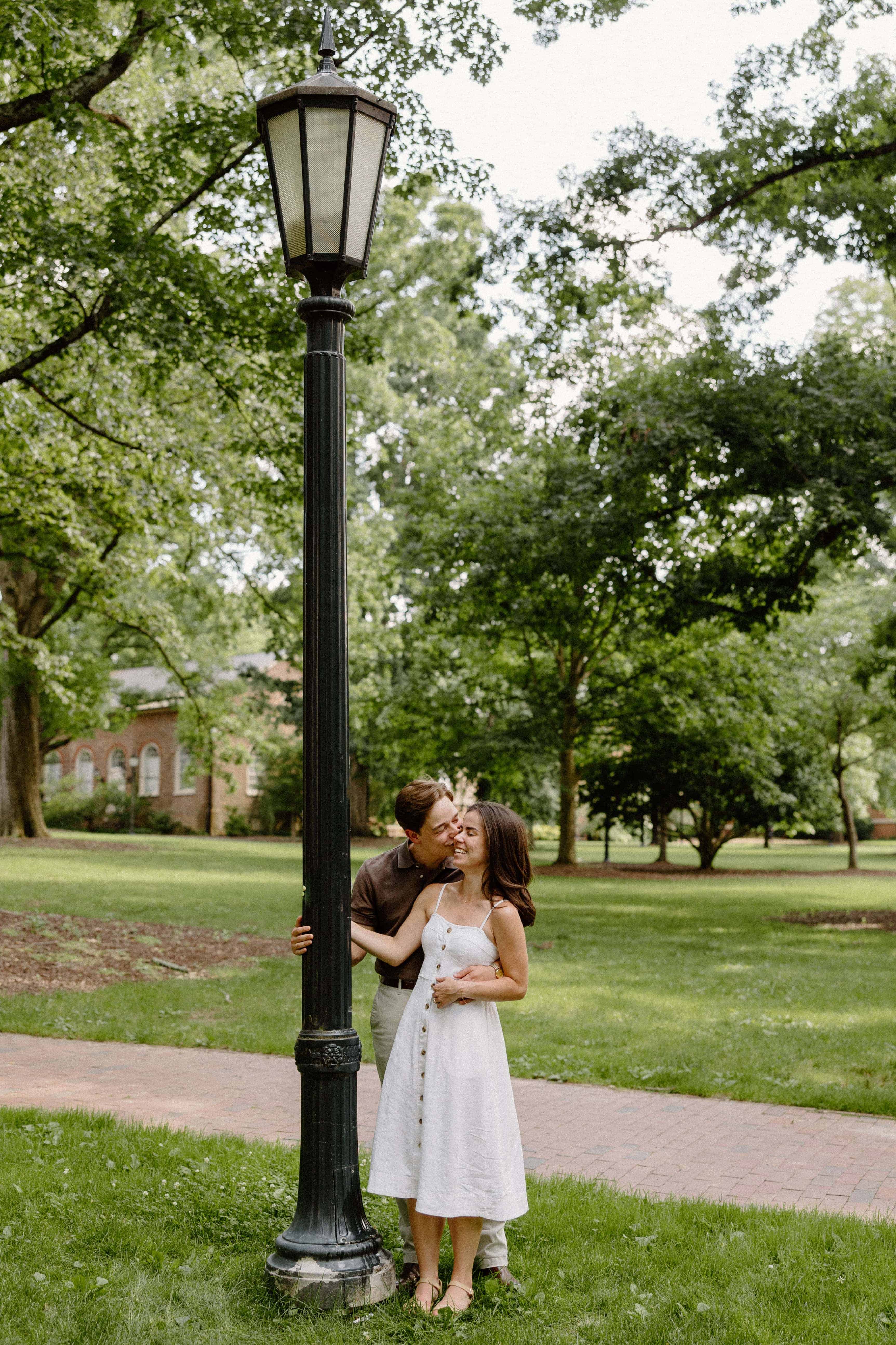 A woman in a white dress is standing on a grassy lawn next to a tall black lamppost. Trees and a brick building are in the background.