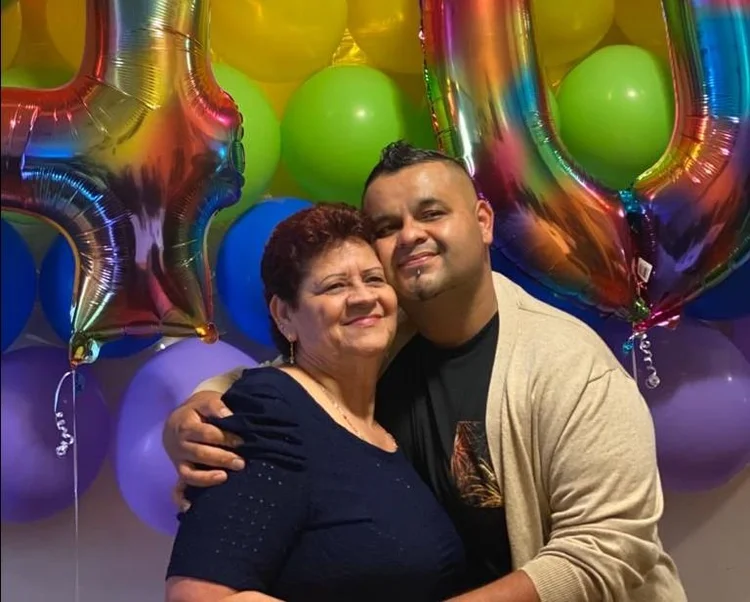 Um homem e uma mulher sorrindo e se abraçando em frente a um cenário colorido de balões, com grandes balões numéricos nas cores do arco-íris, em uma celebração festiva.