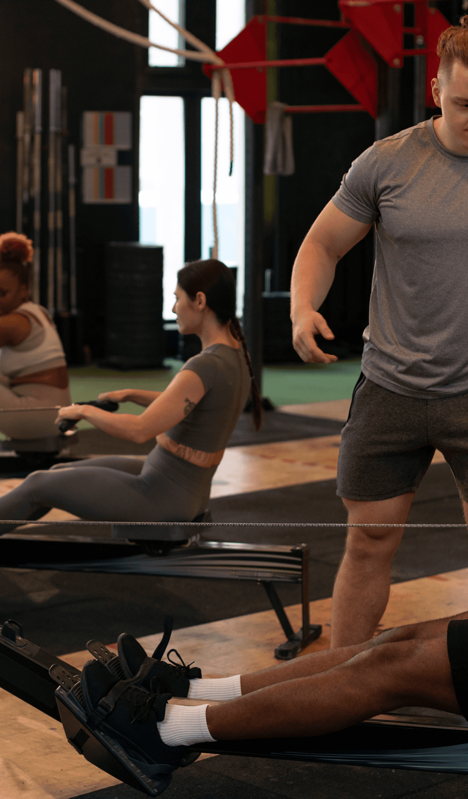 People working out on rowing machines in a gym with a trainer nearby.