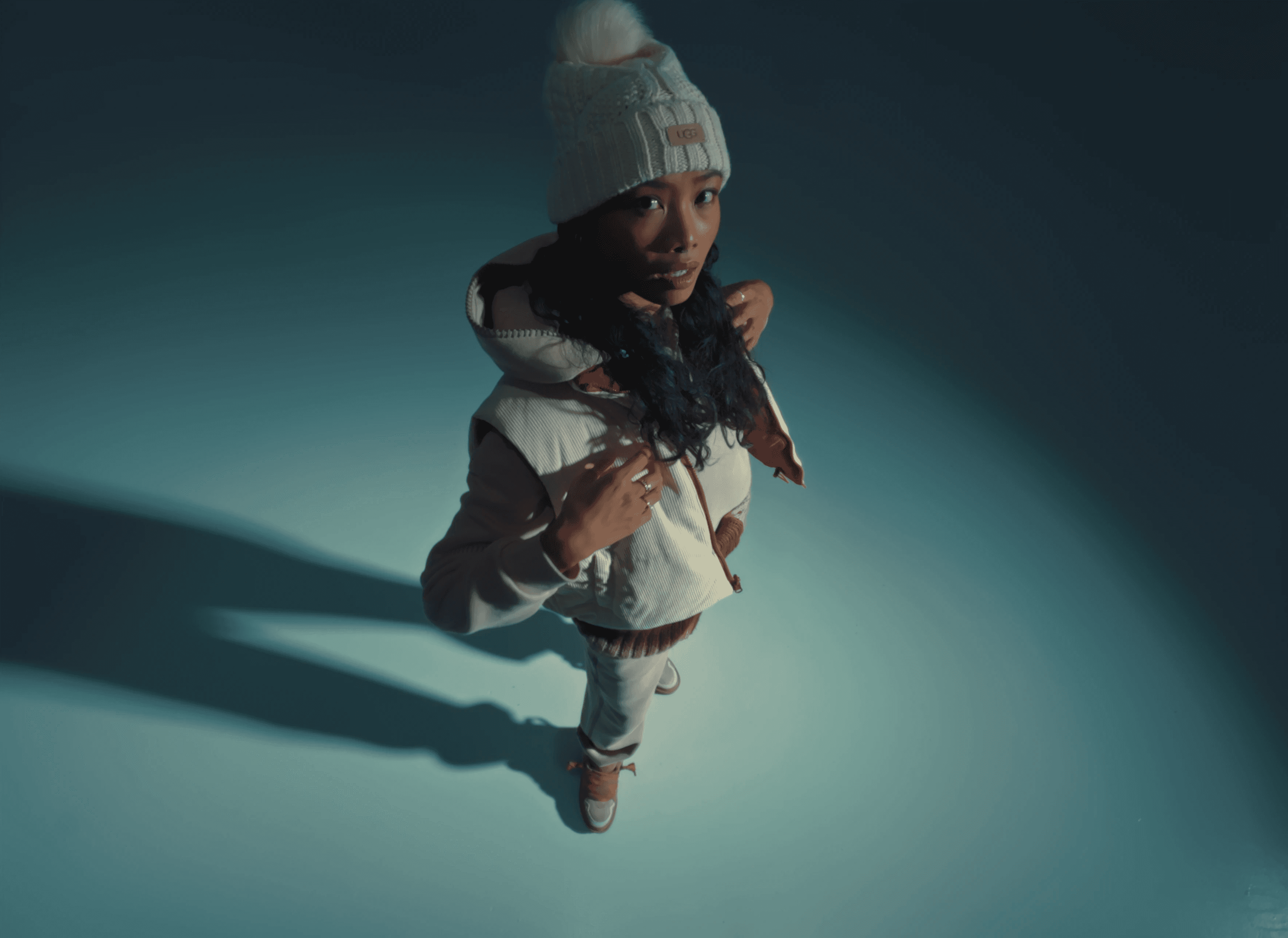 A person dressed in winter attire, including a white knit hat with a pom-pom and a padded jacket, stands on a smooth, light blue surface, casting a long shadow in a dimly lit environment.