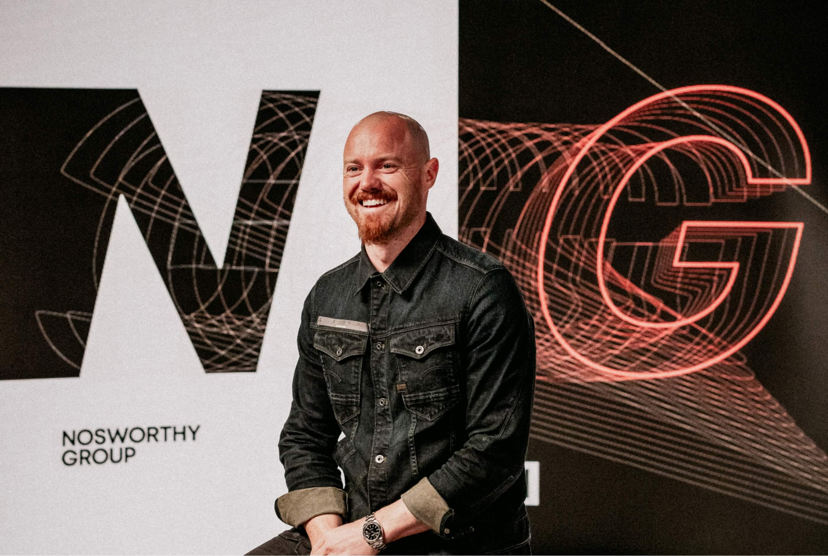 A smiling bald man in a denim jacket seated on stage in front of a screen displaying the Nosworthy Group logo and red line-art "NG" graphics.