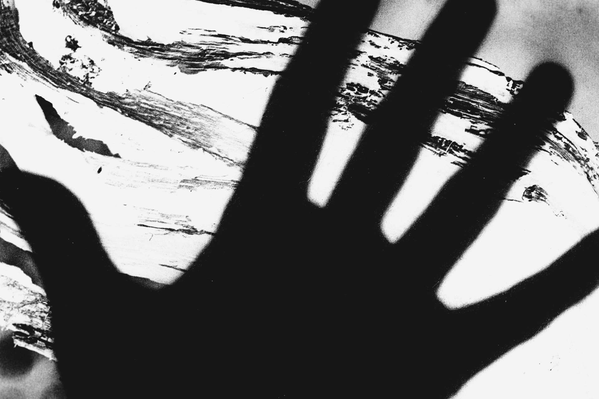 Macro photograph of hand silhouette with spread fingers against layered film negatives showing brush stroke marks, high contrast creating gestural shadow form