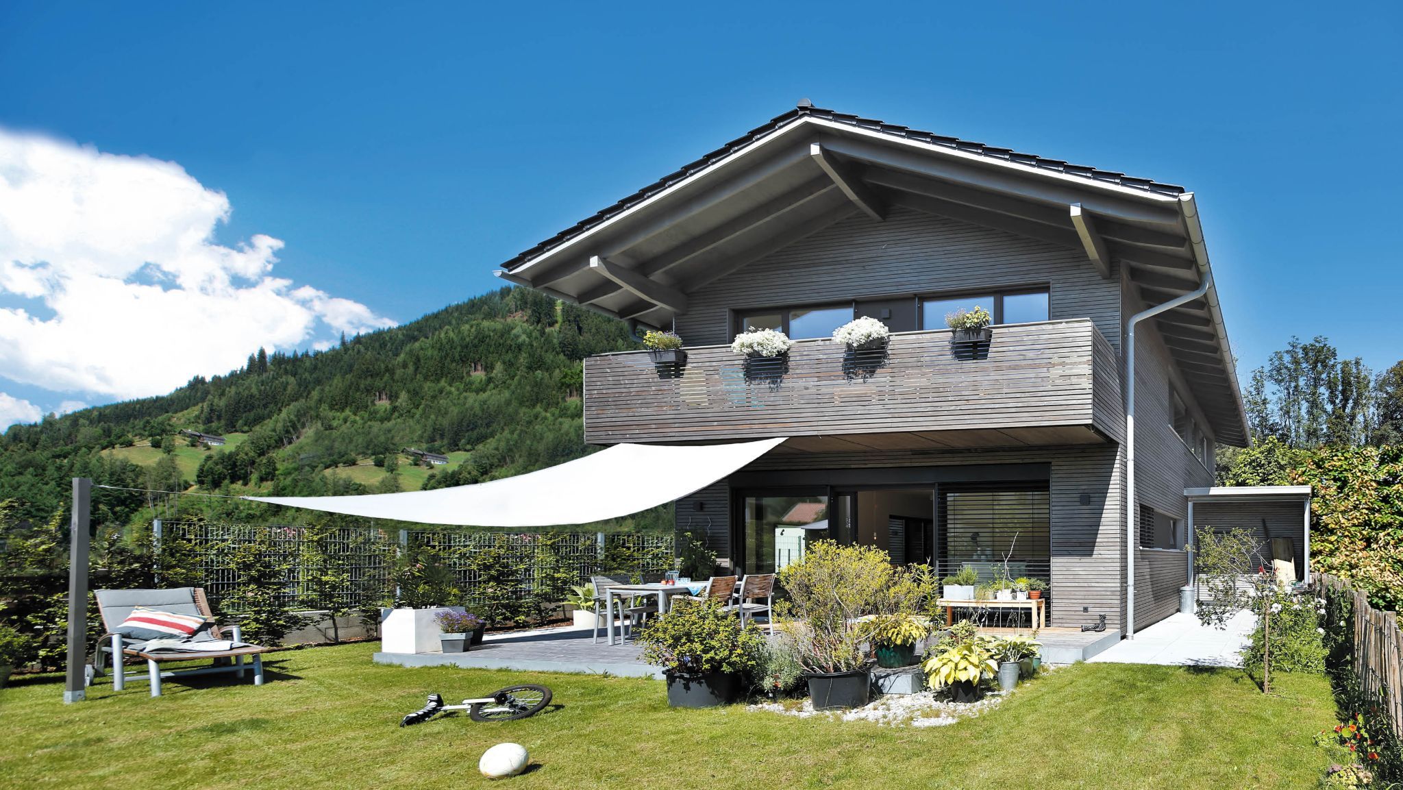 Modernes Holzhaus am Hang mit Terrasse, Sonnensegel und üppigem Garten, umgeben von grünen Bergen.