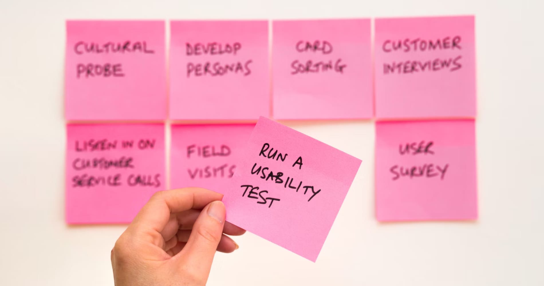 Pink sticky notes on a wall listing UX research methods, with a hand holding one labeled 'Run a Usability Test'