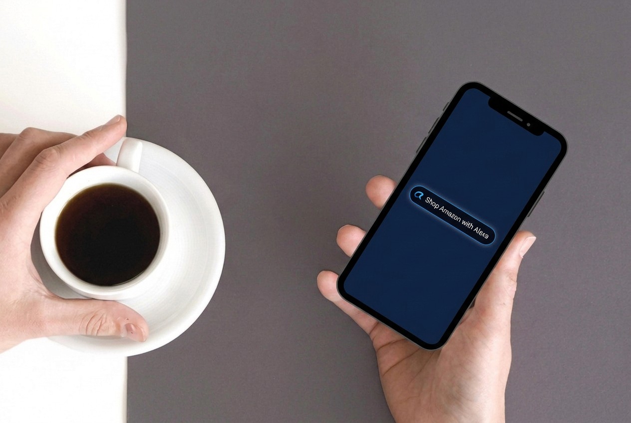 A top-down shot shows two hands interacting with a smartphone and a cup of coffee. The left hand holds a white coffee cup filled with dark coffee, resting on a white saucer. The cup and saucer are positioned on the left side of the frame, on a white surface that transitions to a dark gray surface. The right hand holds a black smartphone, tilted slightly, displaying a search bar with the text "Shop Amazon with Alexa" illuminated in a blue glow. The background is split diagonally, with the left side white and the right side dark gray.
