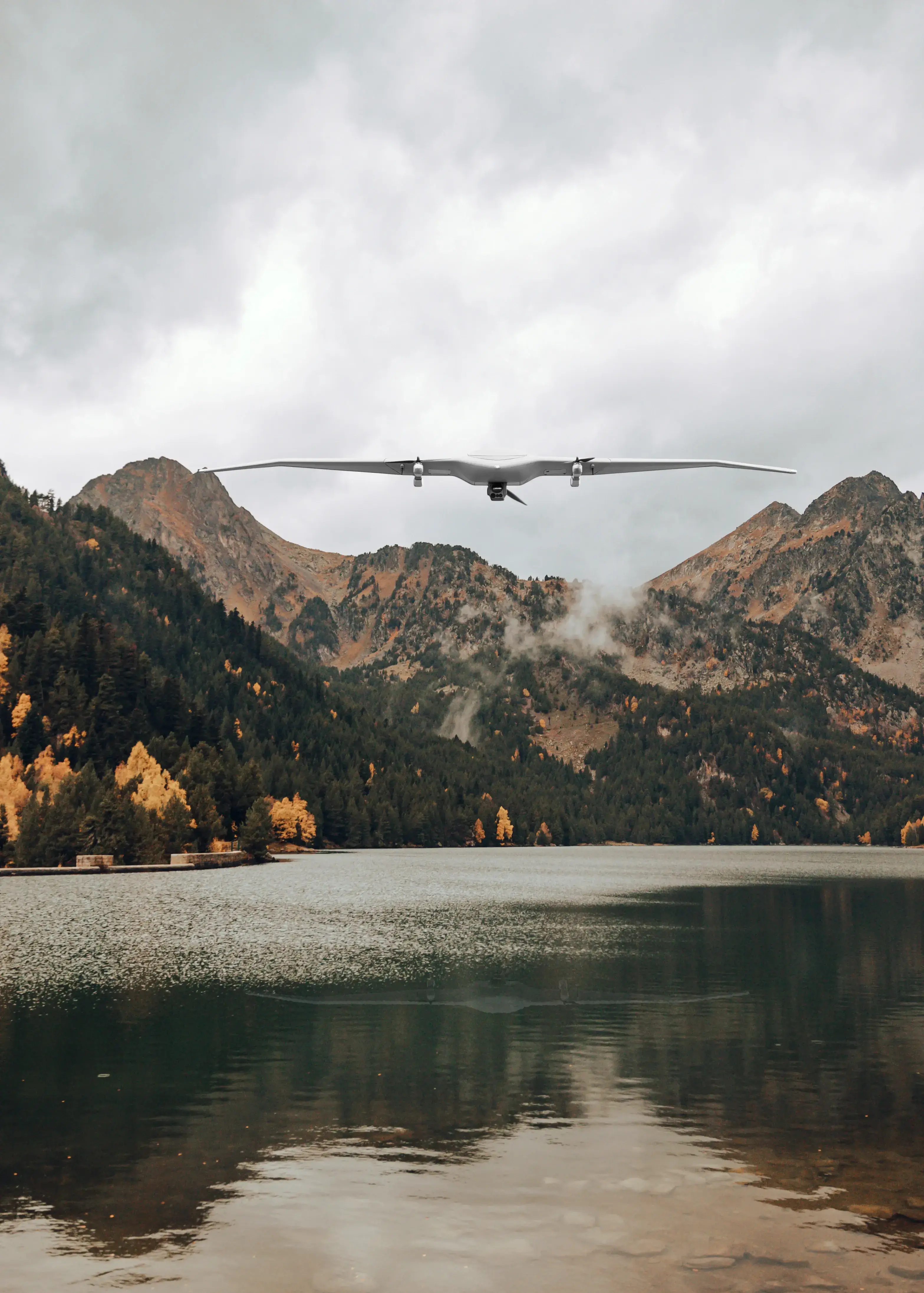 A drone soars over a tranquil lake surrounded by lush, autumn-colored forests and mountainous landscape under a cloudy sky.