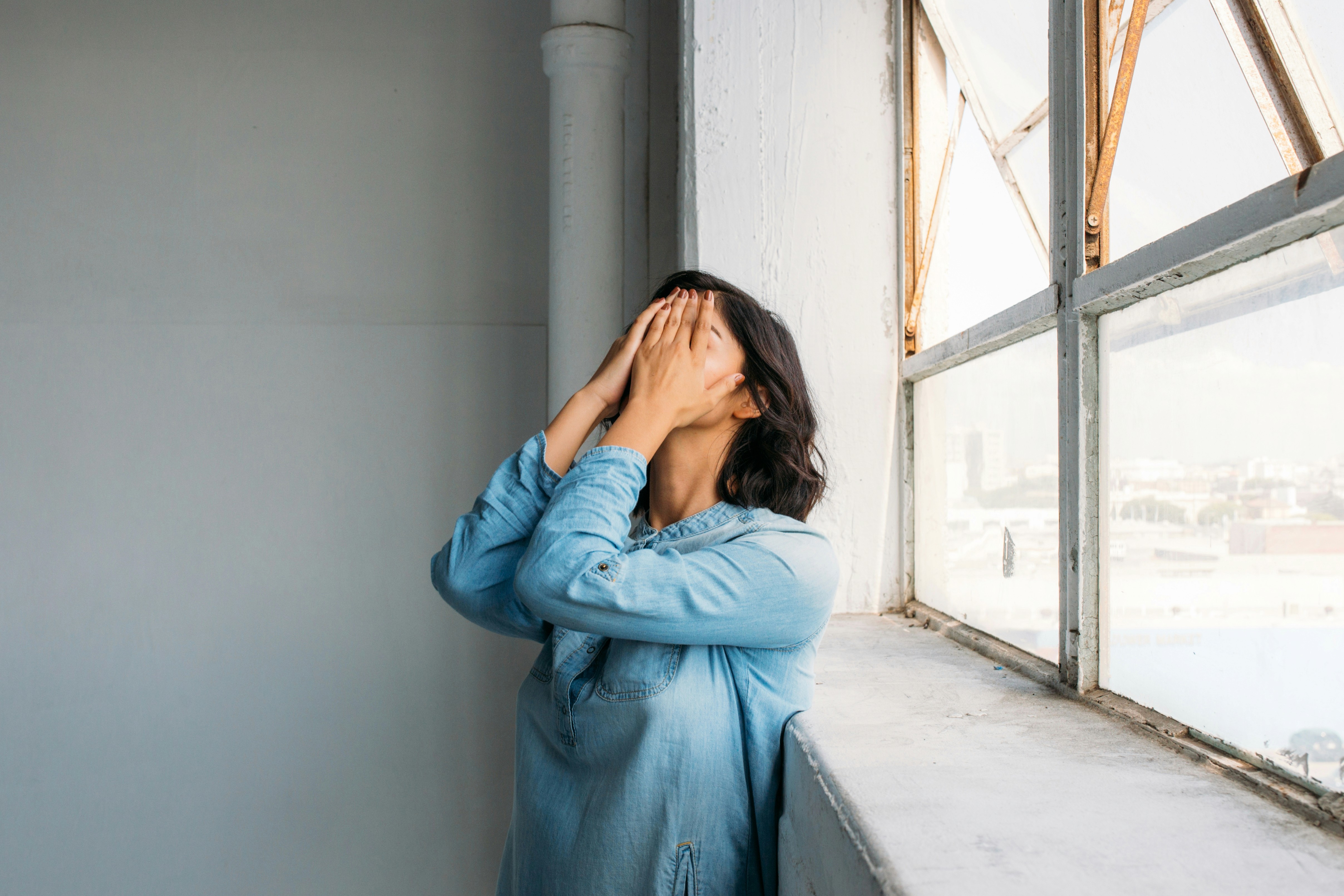 Eine Frau steht am Fenster eines minimalistischen Raumes und bedeckt ihr Gesicht mit den Händen. Das Bild symbolisiert emotionale Belastung, kognitive Überforderung und den Bedarf nach Klarheit und Entlastung.
