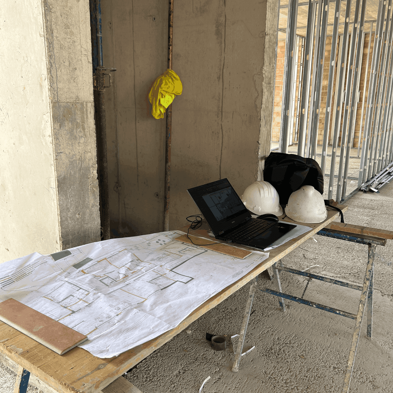 Puesto de trabajo de dirección de obra en un edificio en construcción, con planos técnicos, ordenador portátil y cascos de seguridad sobre una mesa provisional.