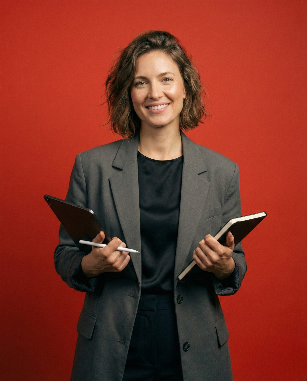 Woman in a gray blazer holding a tablet and notebook, smiling against a red background. Confident and professional tone.