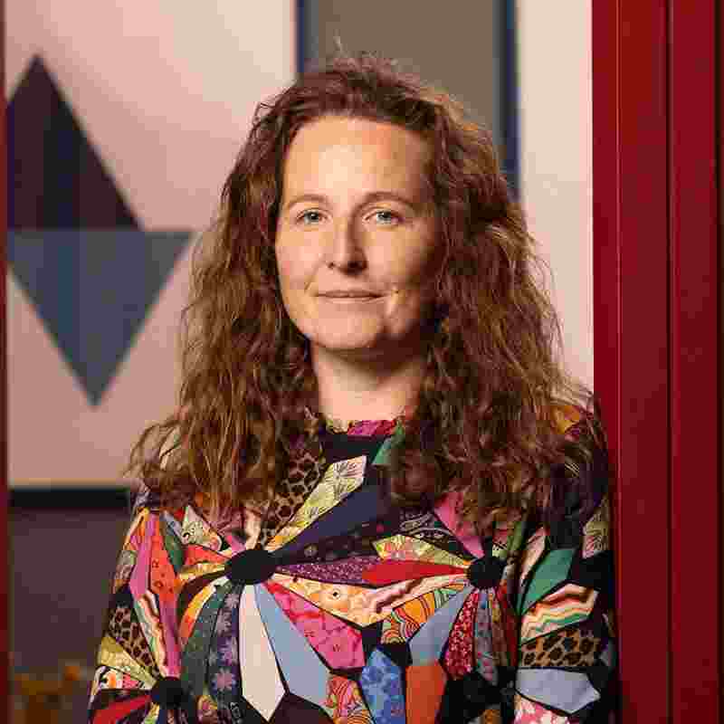 A woman with long wavy brown hair wearing a colourful geometric top smiles lightly at the camera