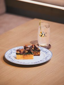 A beautifully plated dish of toast topped with rich, savory ingredients, served alongside a refreshing cocktail in a glass featuring the Roku Gin logo, set on a wooden table.