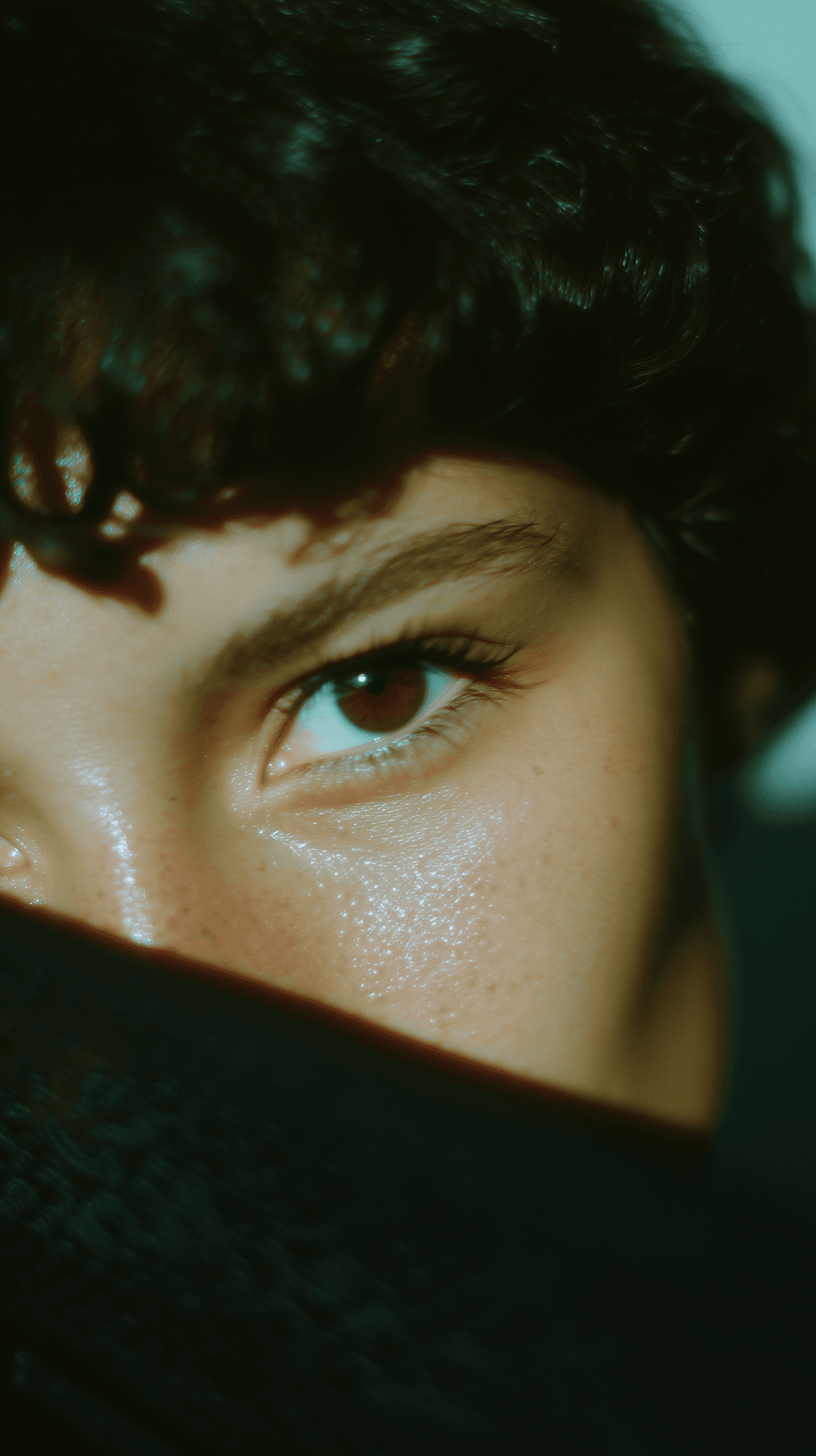 Close-up of a brown eye with dark curly hair and smooth skin, partially covered by a dark fabric, creating a mysterious vibe.