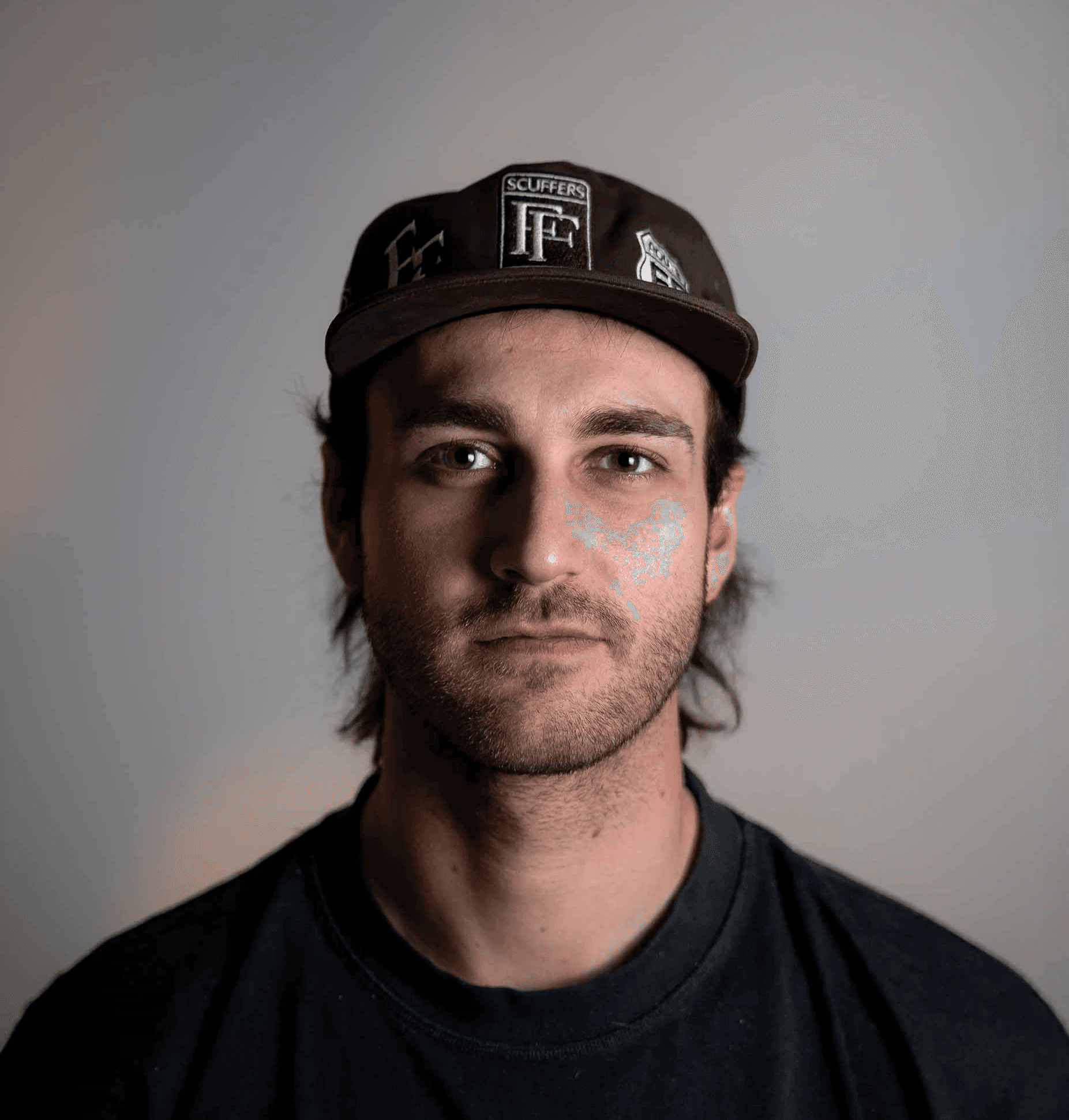 A young man with light brown hair is wearing a black suit jacket and white shirt, standing in a softly lit office setting, conveying professionalism.