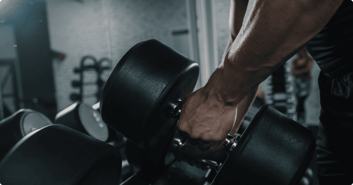 A close-up of a person's hand gripping a large dumbbell, ready to lift it from a weight rack. The gym setting is dimly lit, highlighting the muscular definition in the individual's arm and the heaviness of the dumbbells. 