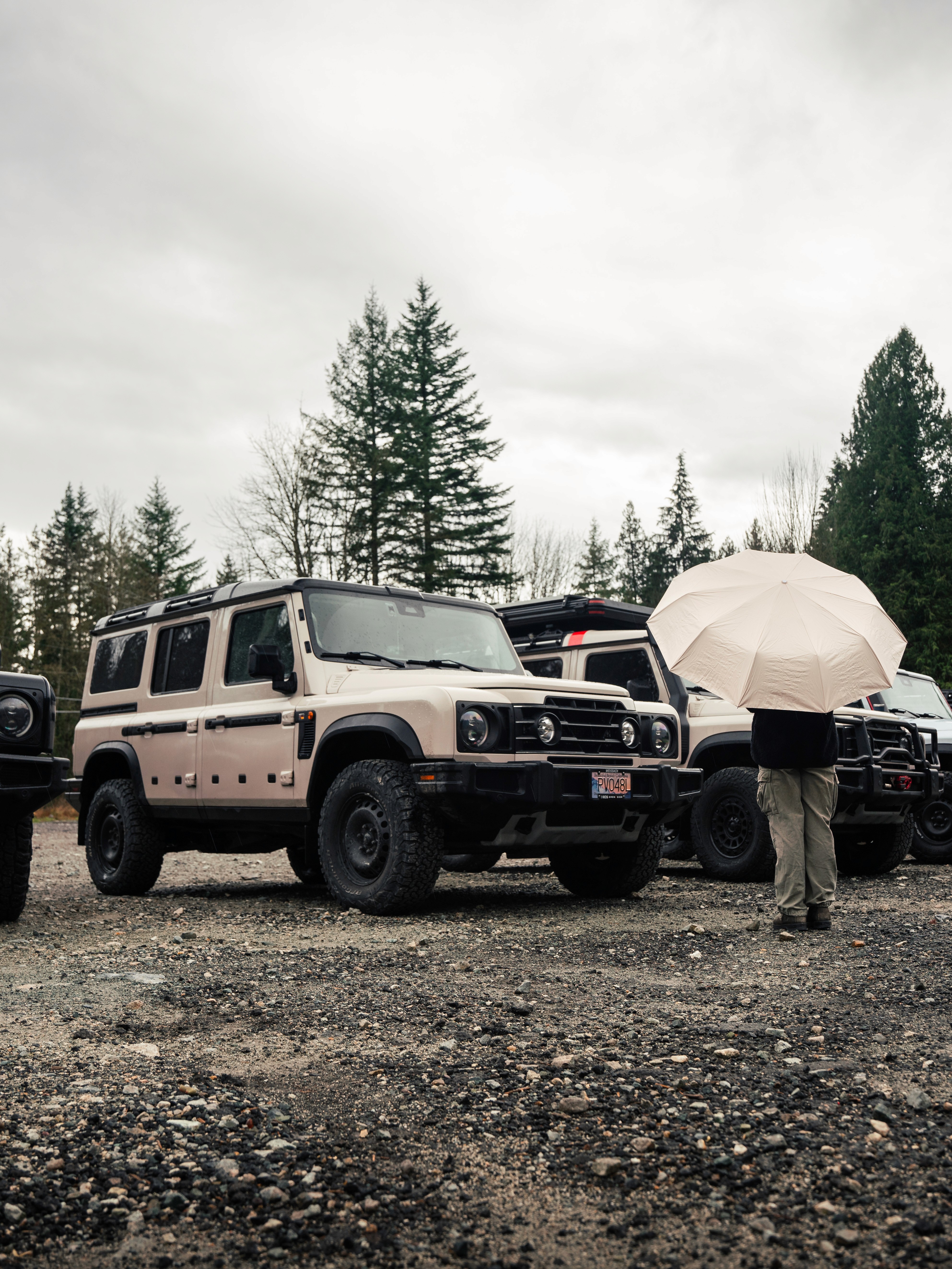 Beige INEOS Grenadier parked on gravel in rainy weather with off-road modifications and overlanding vehicles in the background.