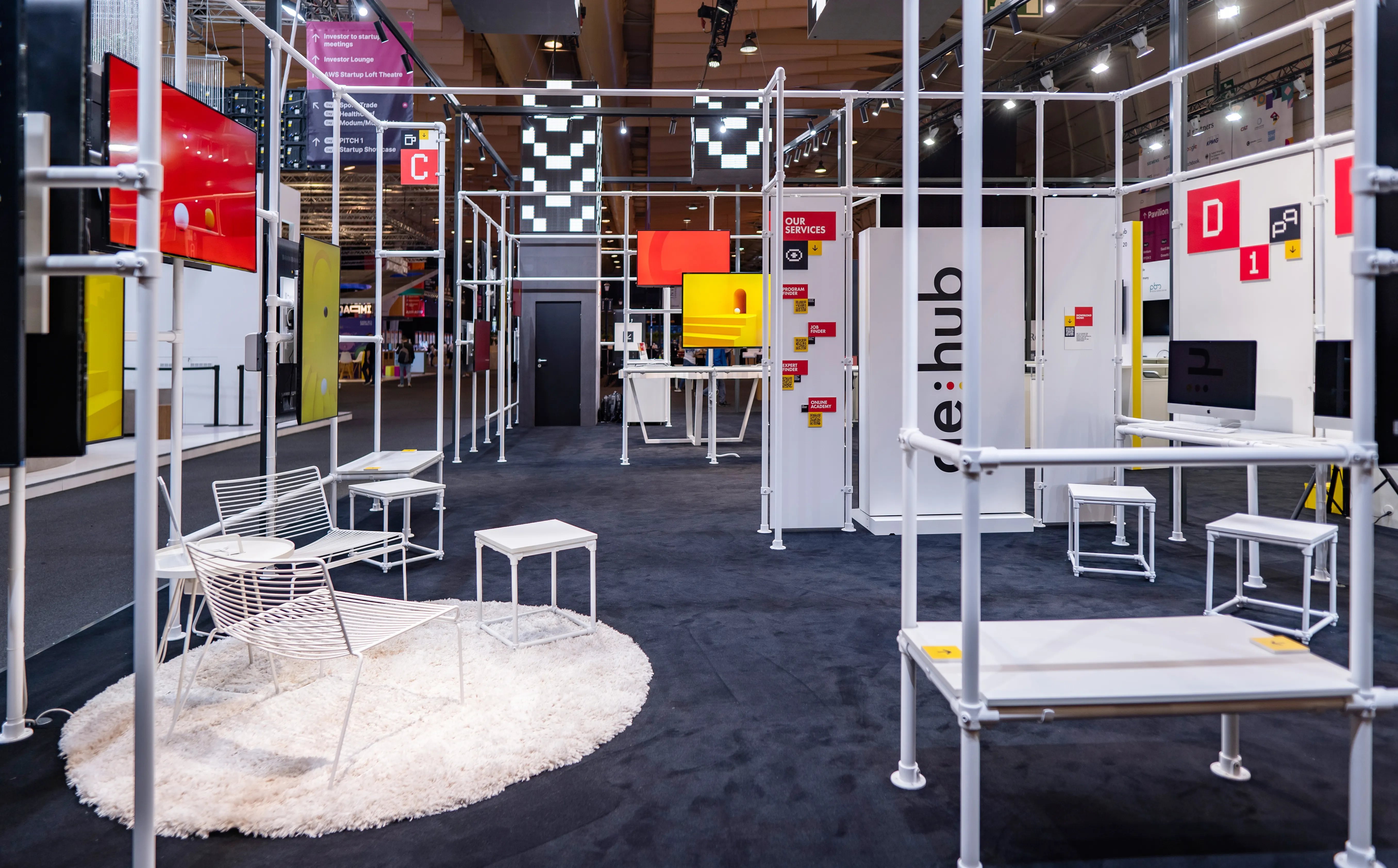 Open booth architecture of the German Pavilion at Web Summit with white tube framework, wire chairs and colorful display panels.