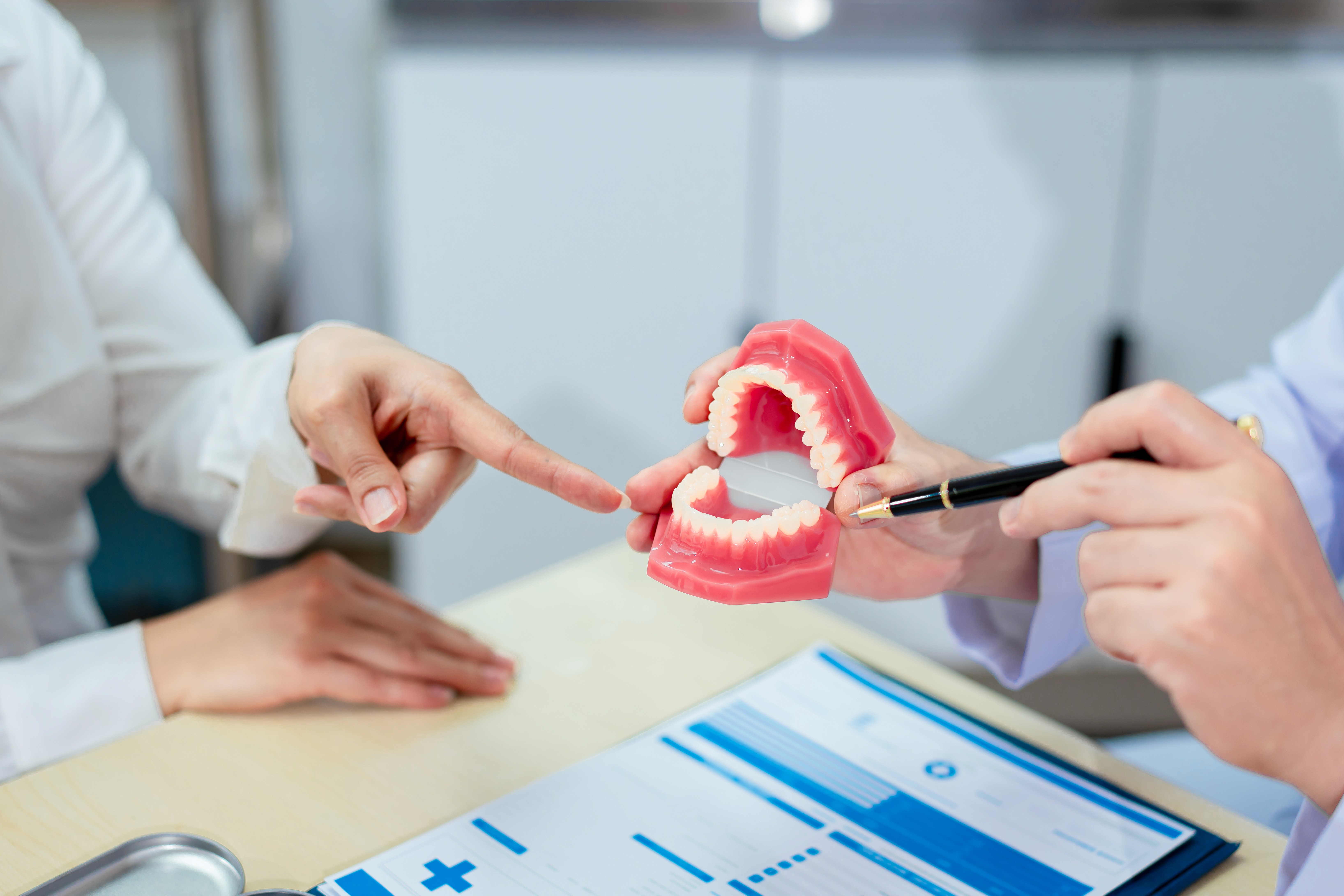 n Asian male dentist explains dental health to a middle-aged Asian female patient while holding a dental model, demonstrating oral care and treatment procedures in a clinical consultation