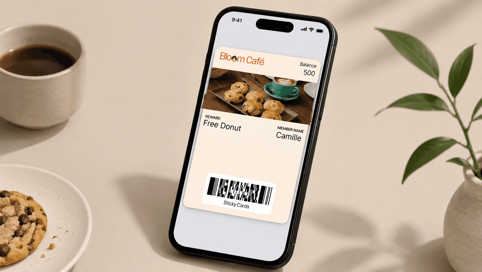 A realistic editorial-style scene featuring a modern smartphone standing at a slight angle on a warm cream surface. Displayed on the screen is a digital “Bloom Café” loyalty wallet pass card with a photo of pastries and a latte, reward details, a member name, balance points, and a barcode at the bottom. Surrounding the phone are subtle café-inspired elements including a cup of coffee, a chocolate chip cookie on a plate, and a small green plant in a ceramic vase. The composition uses soft natural lighting, warm neutral tones, and a clean, premium aesthetic that blends café culture with modern mobile loyalty technology.