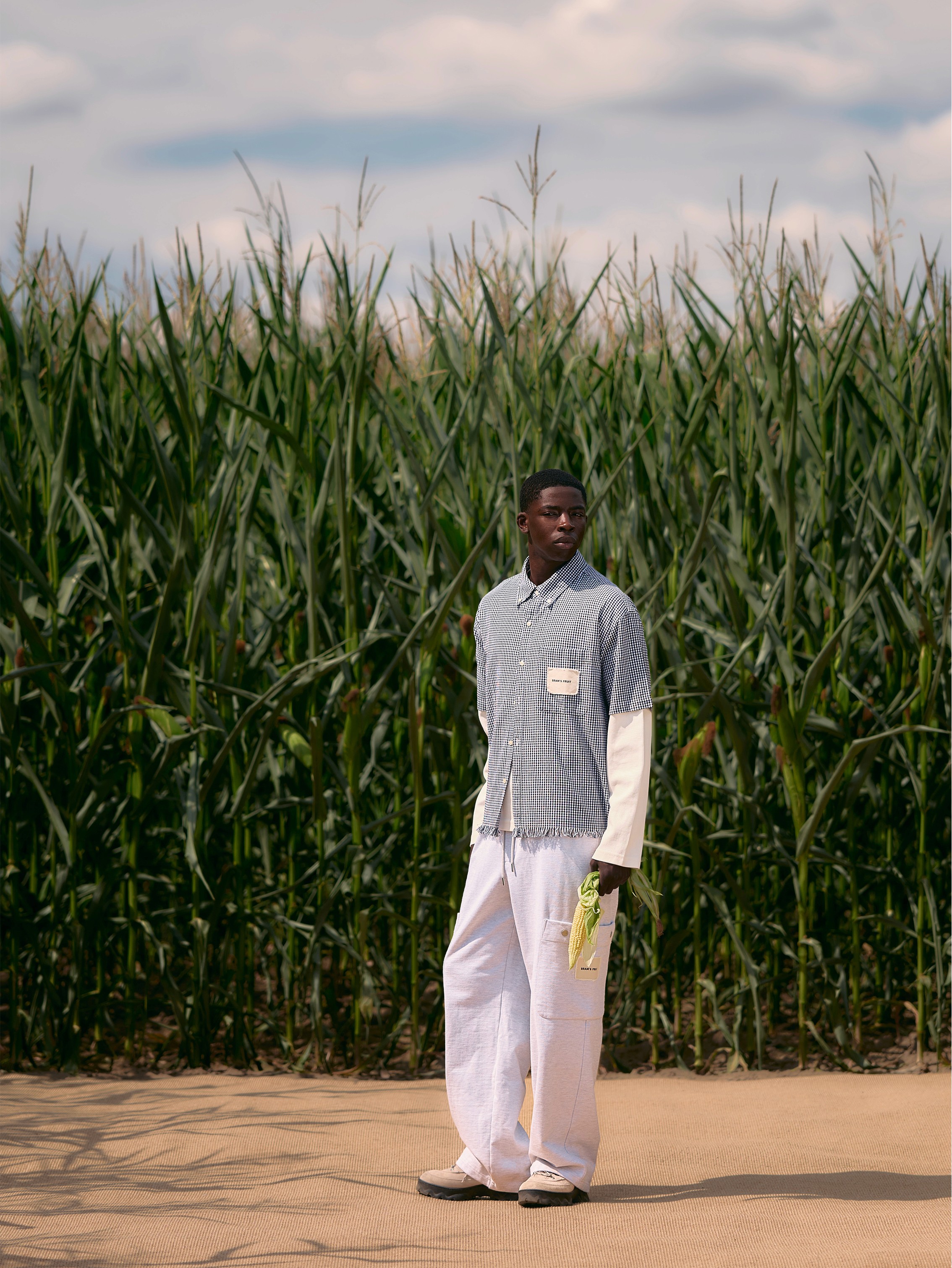 Devant un champ de mais, le mannequin porte un pantalonblanc et une chemise à petits carreaux effilochés de la marque Bram's Fruit