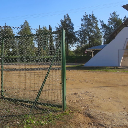 在晴朗的蓝天下，一个土地区域内有一扇绿色铁丝网门，背景中有树木和一座白色建筑。