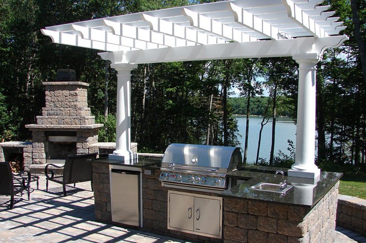 Outdoor kitchen under white pergola overlooking lake and trees