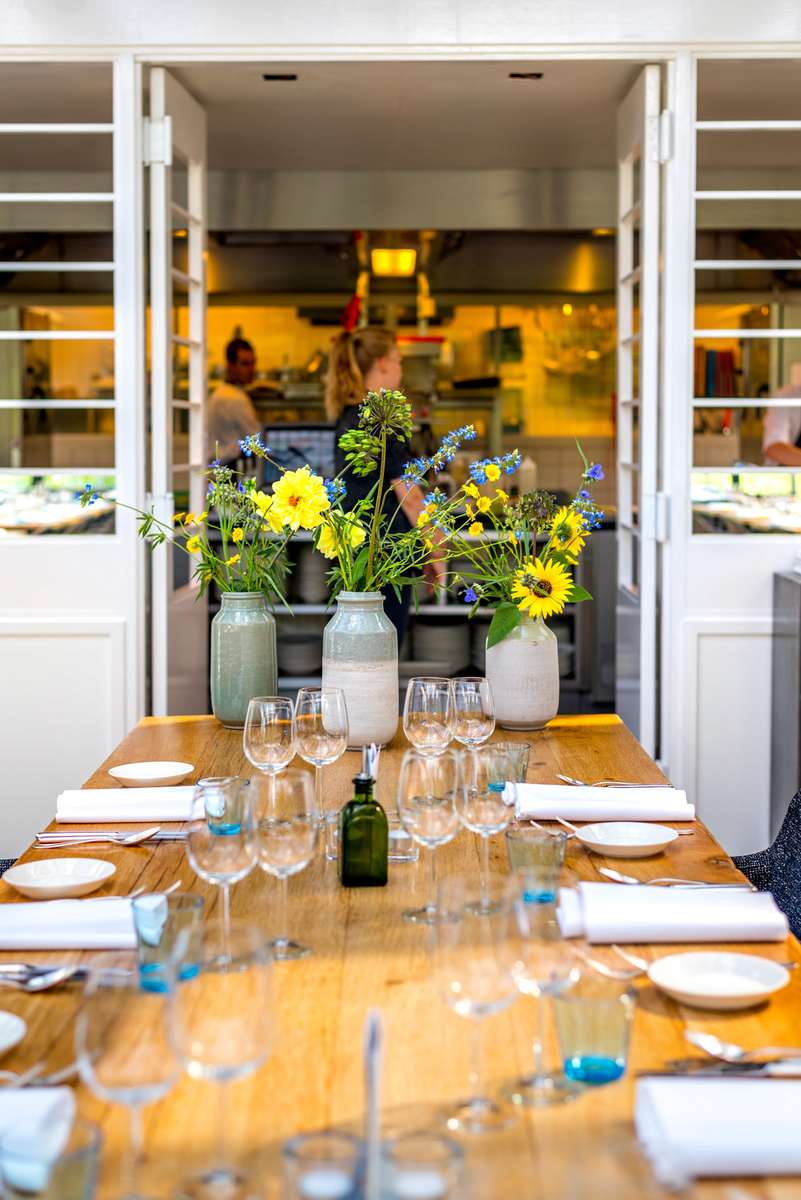 A long wooden table set with plates and glassware in front of an open kitchen door decorated with yellow flowers.