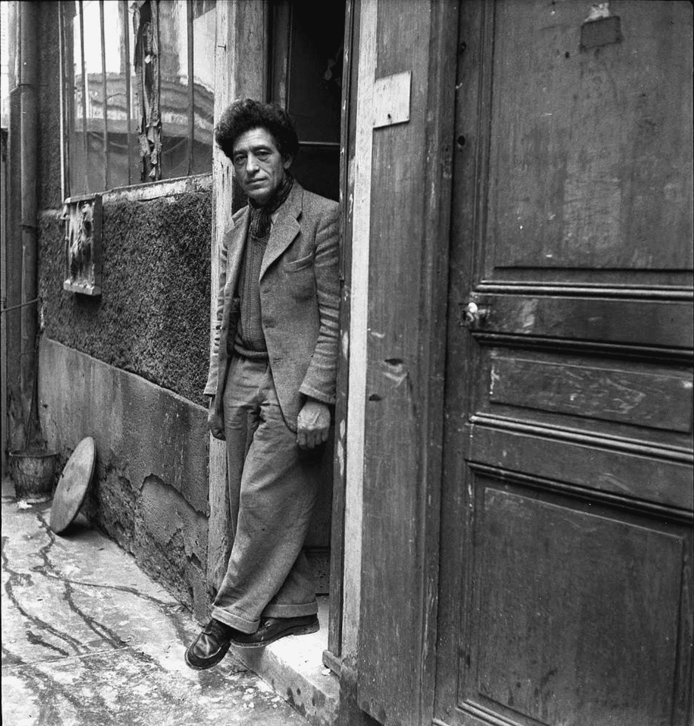 Alberto Giacometti standing in the doorway of his Paris studio courtyard, black and white historical photo for Giacometti Museum & School in Paris