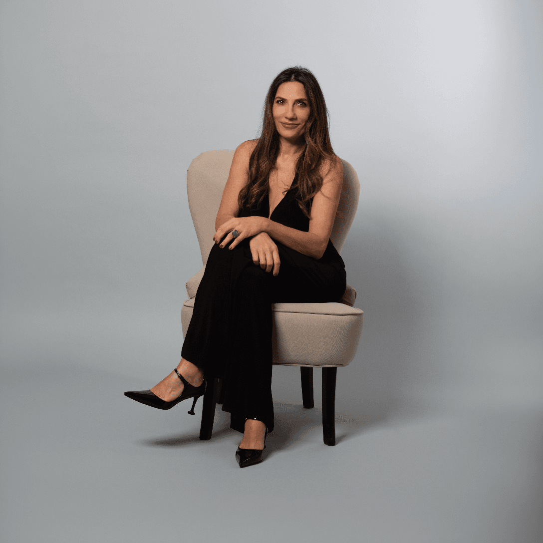 A young person with curly hair smiles at the camera while sitting in front of a laptop against a dark background.