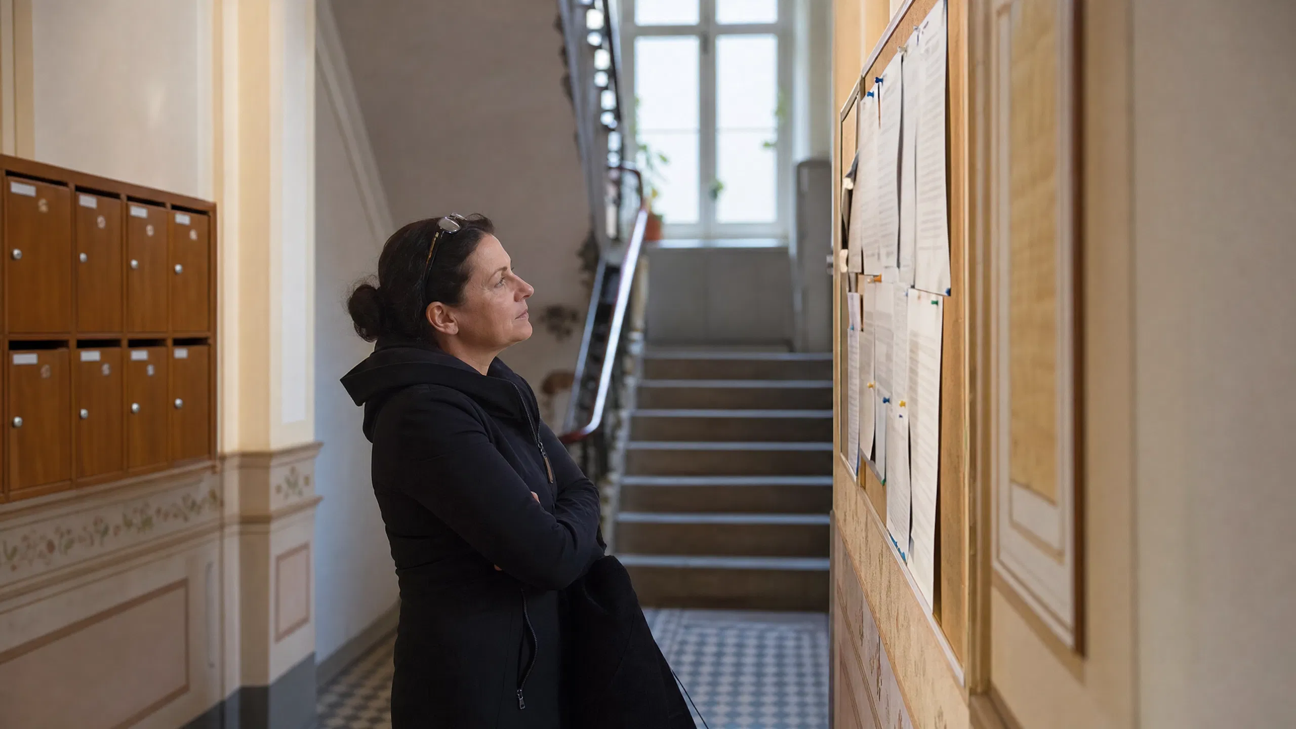 Eine Bewohnerin mittleren Alters steht im Eingangsbereich eines Wiener Altbaus und liest aufmerksam die Aushänge am Schwarzen Brett – links die Holzbriefkästen, im Hintergrund das geschwungene Stiegenhaus mit Tageslicht von oben.