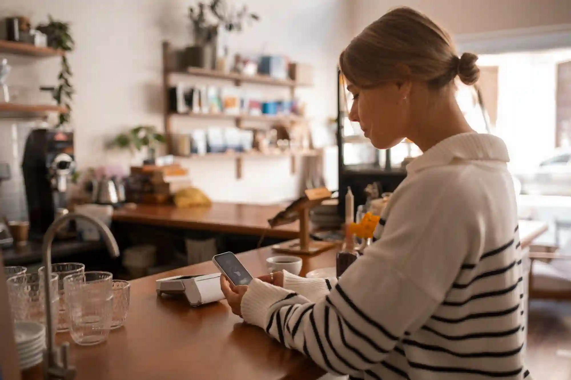 A woman in a striped shirt checks her phone at a cafe counter, managing her social campaign.