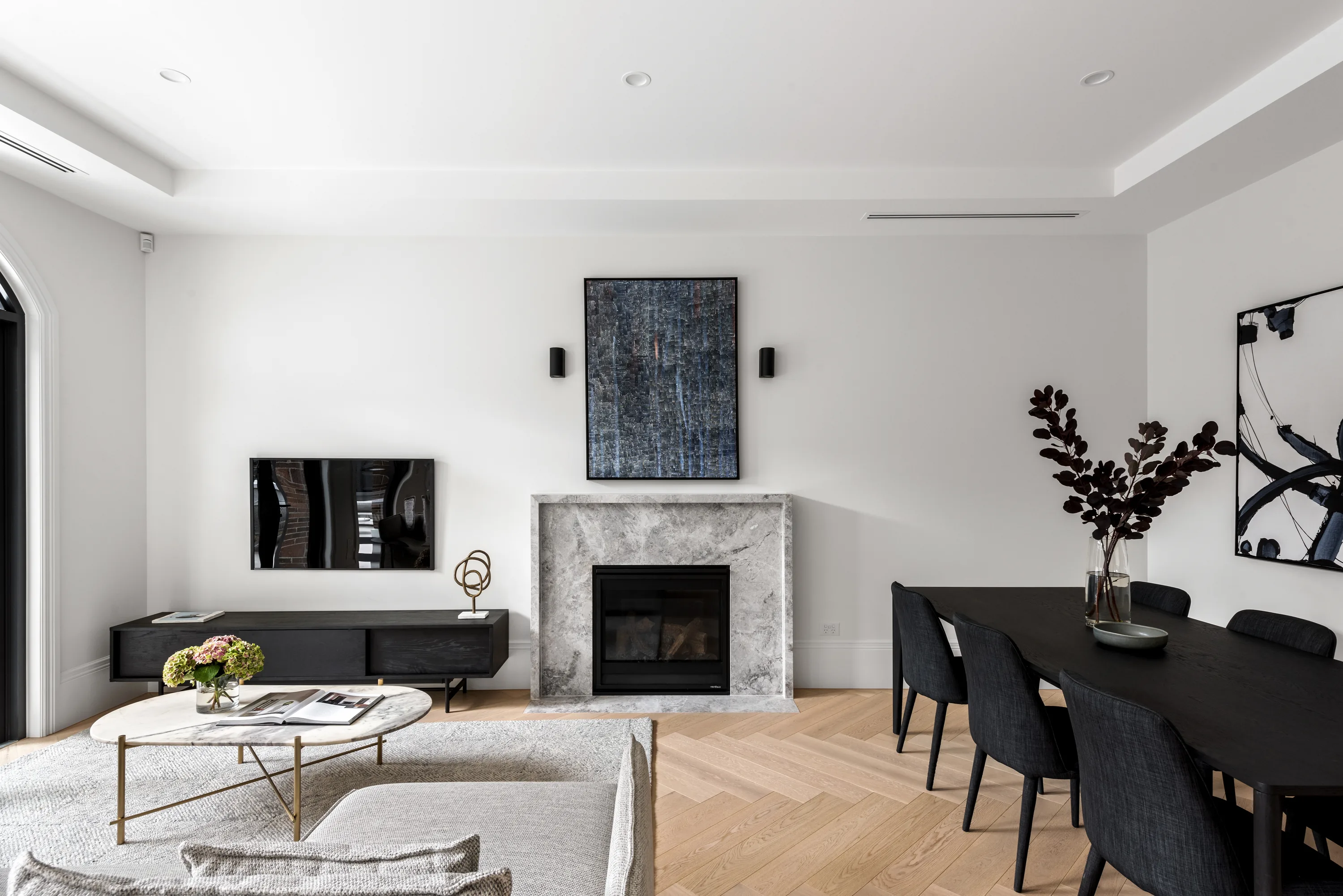 Living and dining area viewed toward an arched doorway, with a grey-marble fireplace, framed art, pale sofa, marble coffee table and a black dining setting on timber floors