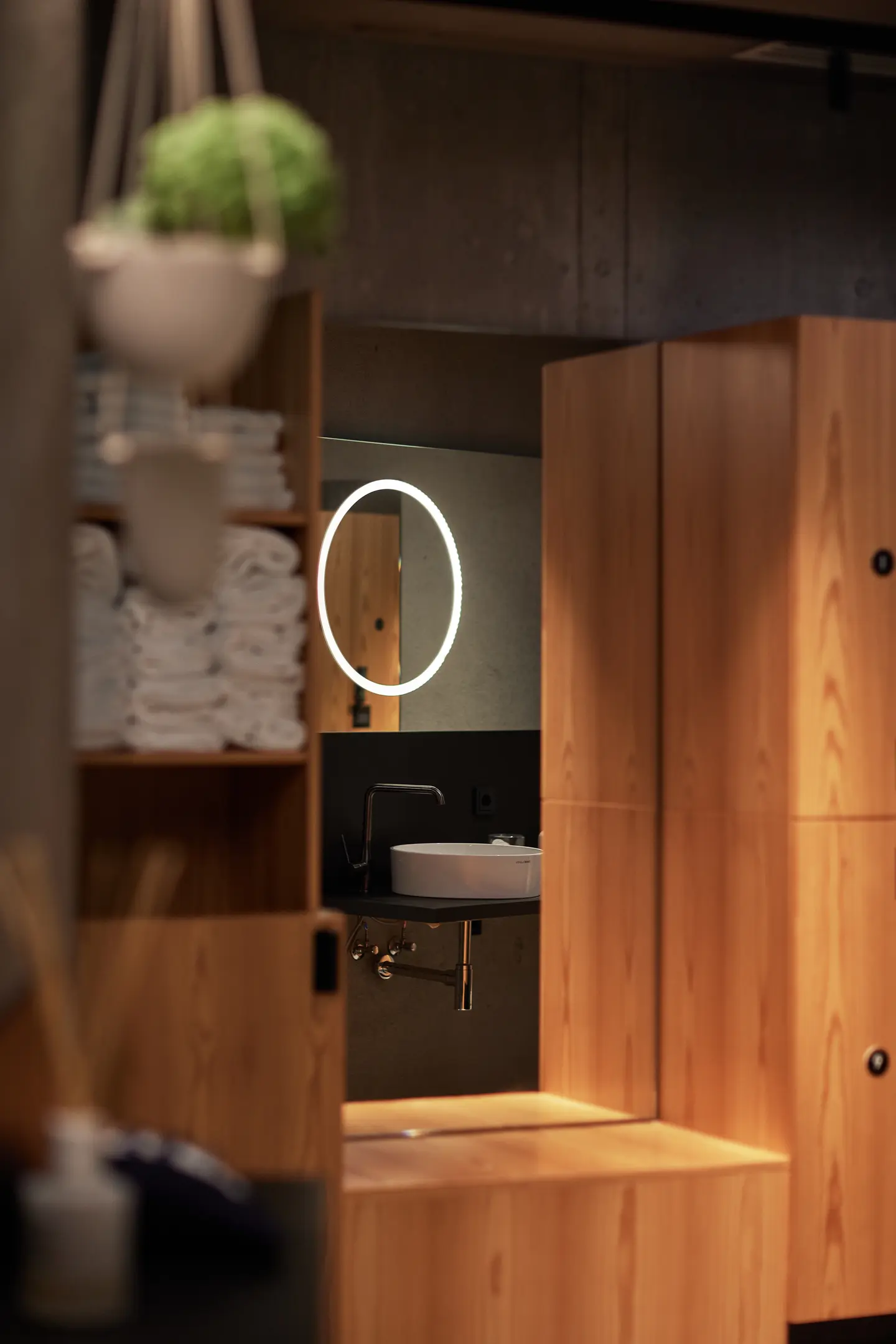 Wooden sauna changing area with a round illuminated mirror, sink, towels, and hanging plant