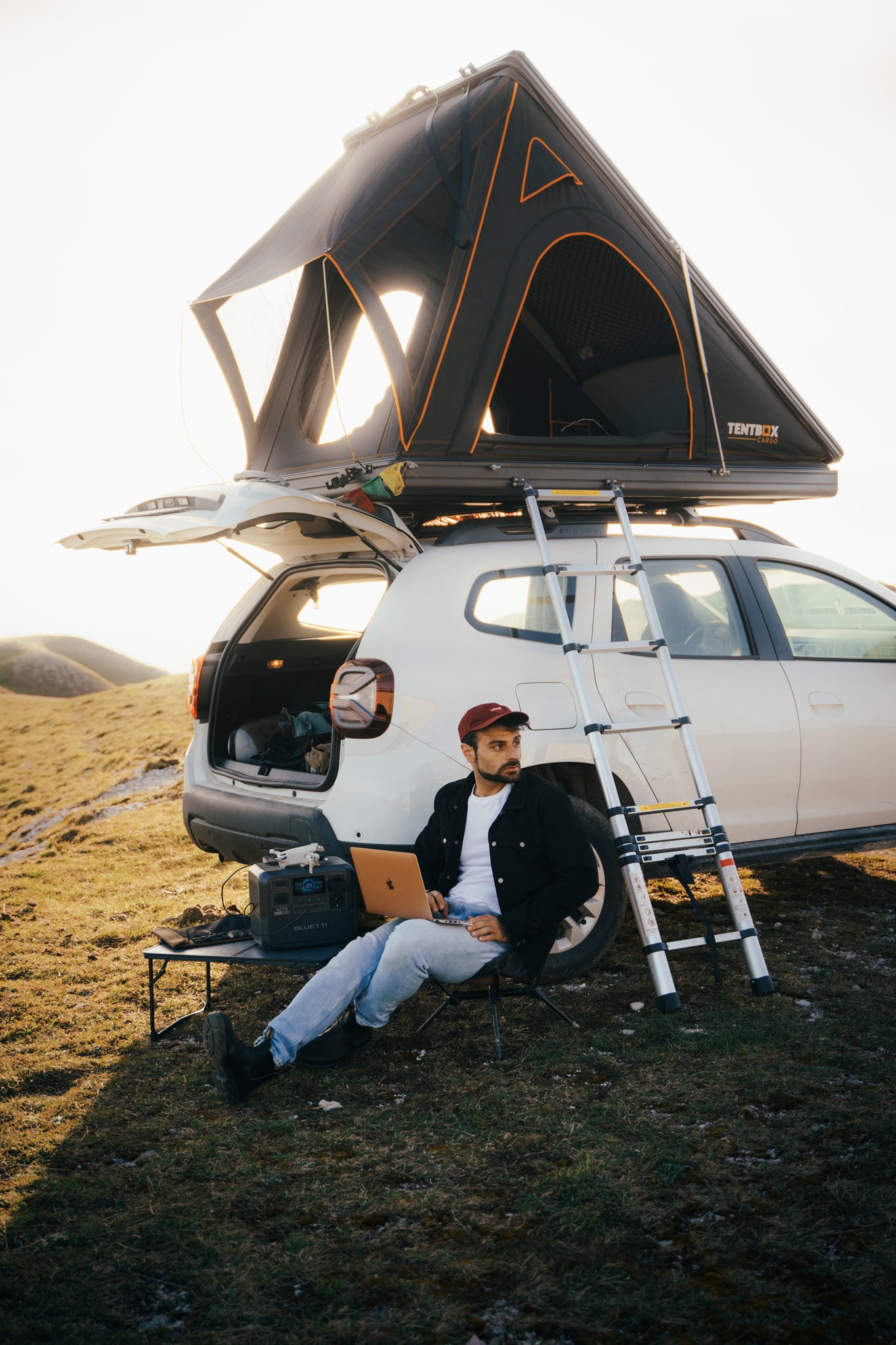 videomaker editing with a pc on his lap in front a car with a rooftop tent