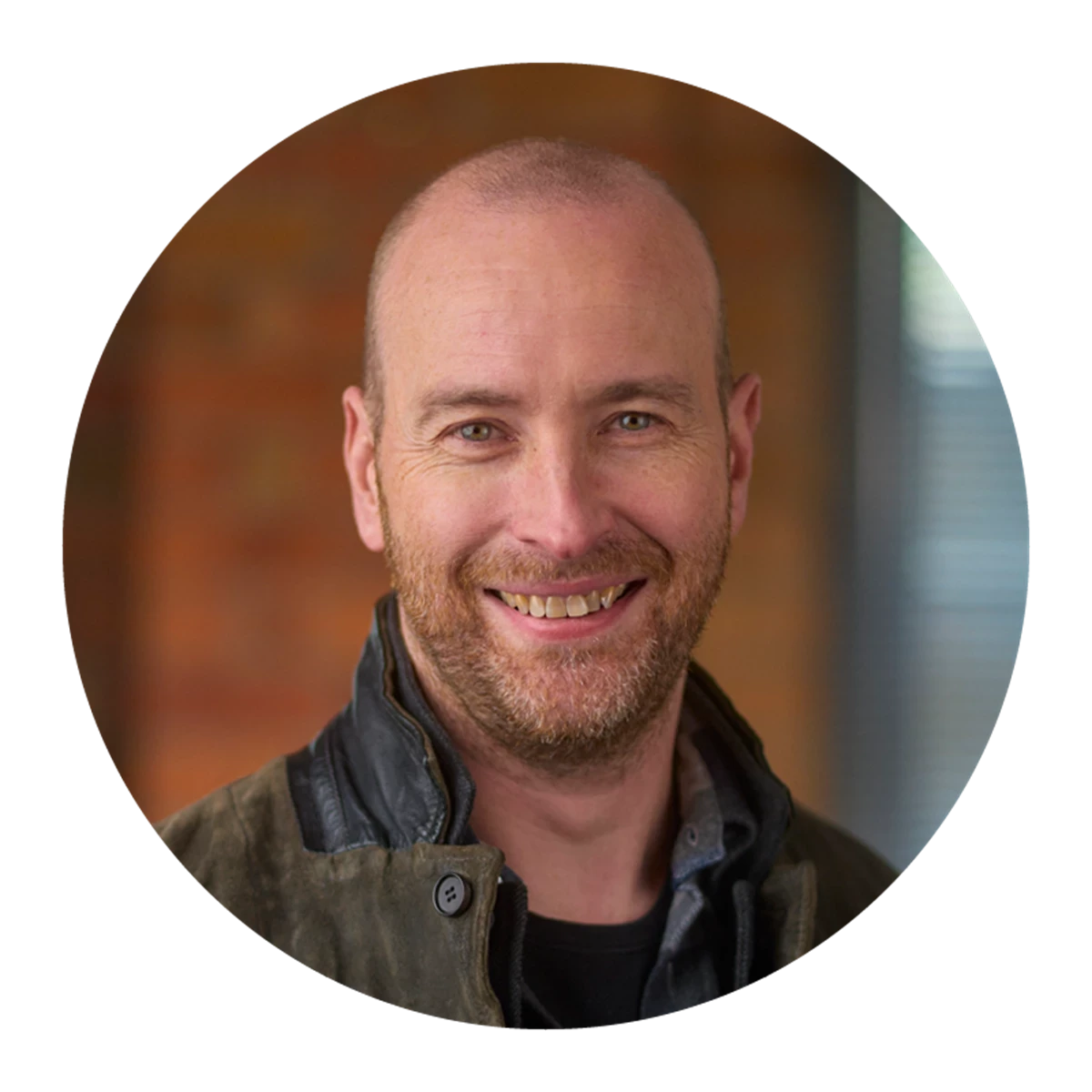 A circular portrait shows a person with closely cropped hair wearing a dark jacket over a T‑shirt. A softly lit indoor background with warm textures is visible.