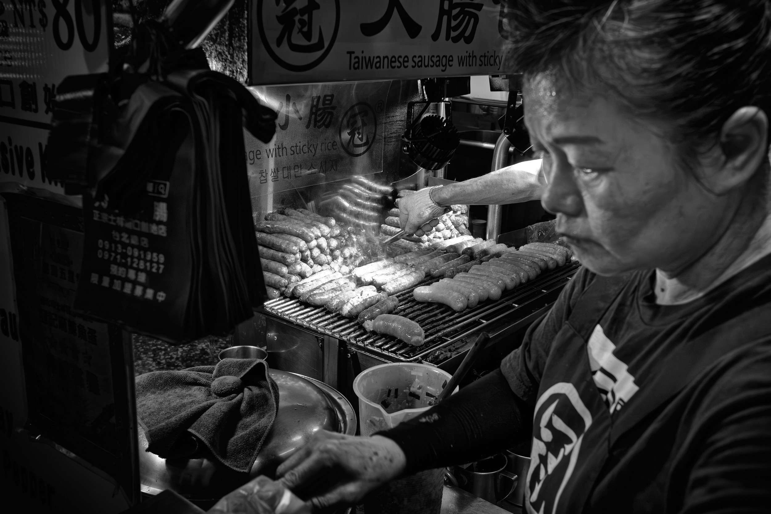 Black and white photograph of two people grilling sausages at a busy night stall with smoke rising off the grill.