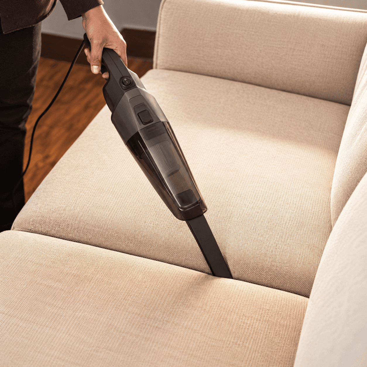 A hand guides a handheld vacuum nozzle along the seam of a beige fabric sofa.