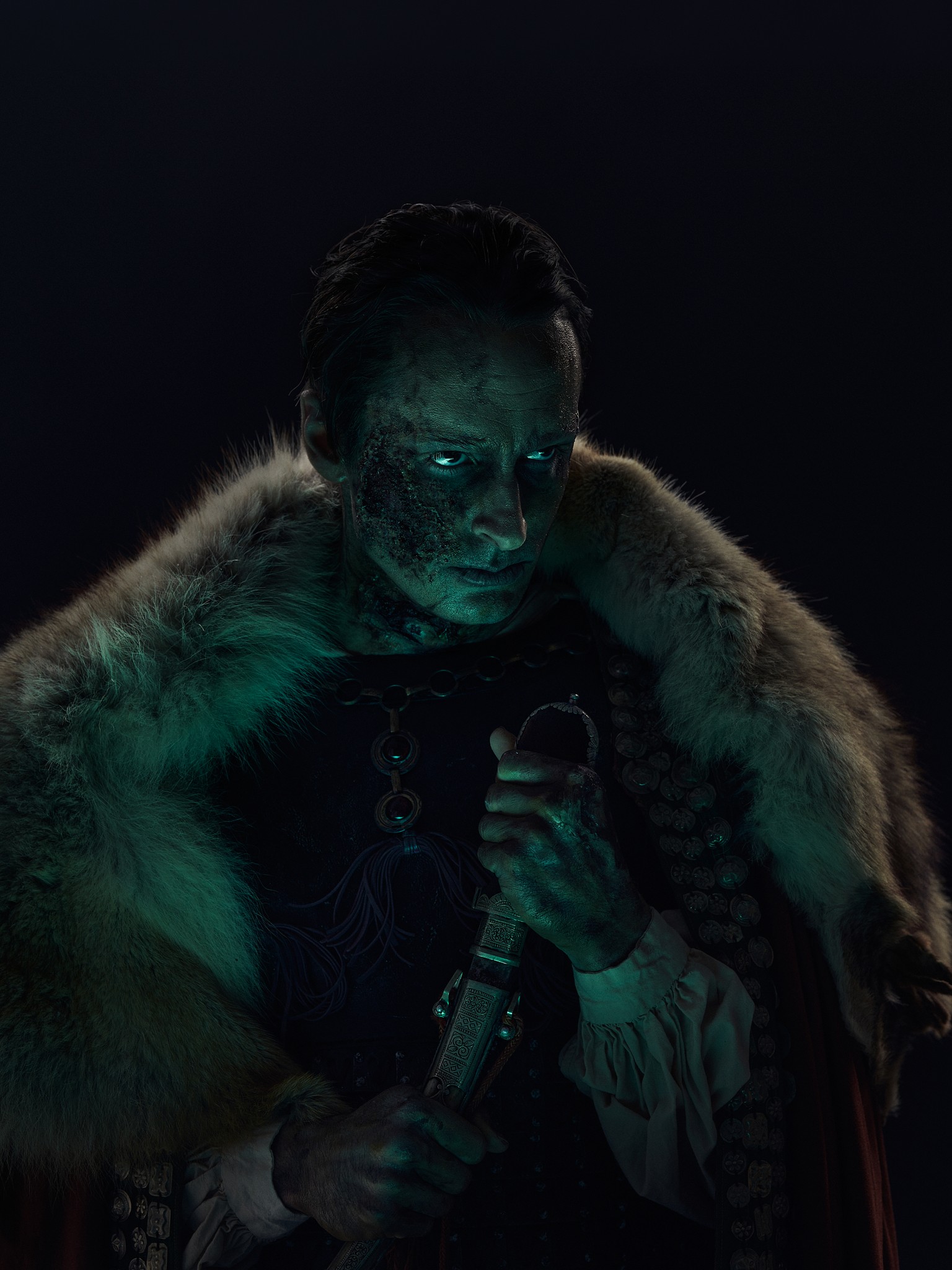 A dark and sinister character portrait of a historical fantasy warrior. The photograph is in the style of film and television key art, publicity images. Shot in the studio in New Orleans, Louisiana. The lighting is technical and moody with green tones the help produce an ominous and foreboding feel. The character is wearing armour, furs and welds an ancient knife. 