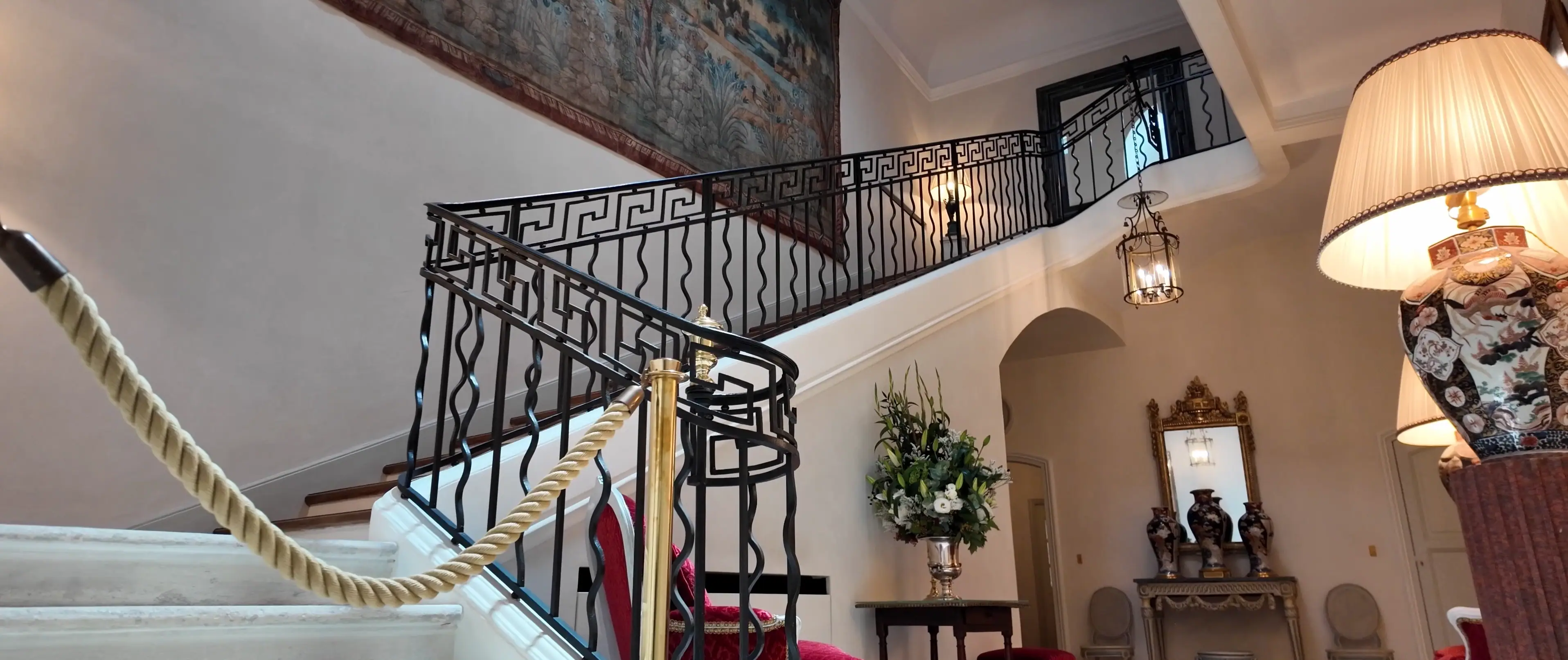 This elegant interior showcases a grand staircase with intricate wrought-iron railings, leading up to a softly lit landing beneath a high ceiling, adorned with a large tapestry, while a gold rope subtly guides the path against a backdrop of classic decor.
