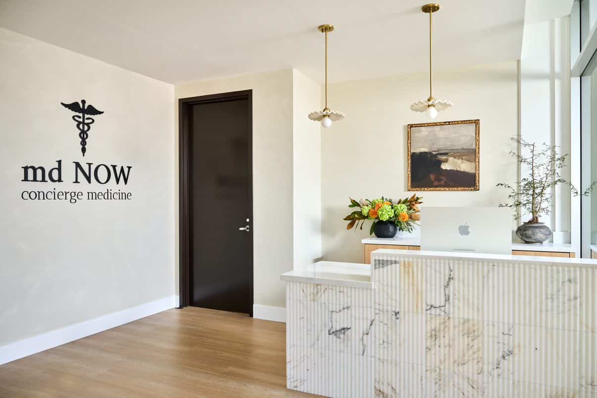 Dimensional “md NOW concierge medicine” wall sign in a bright medical clinic reception area with marble front desk and modern lighting.