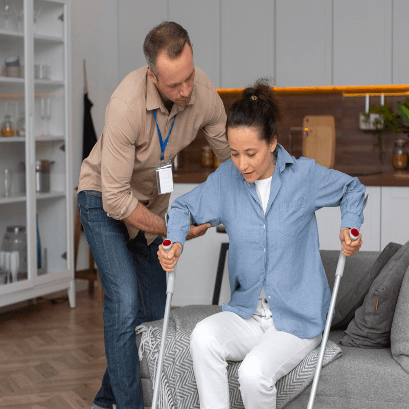 A man assists a woman using crutches as she stands up from a sofa in a modern living room, suggesting a supportive or caregiving situation.