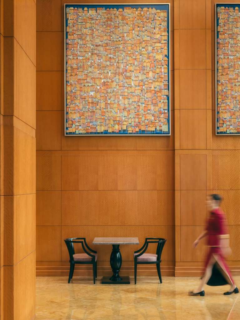 A modern indoor space featuring a large colorful abstract painting on the wall, a small table with two black chairs, and a blurred figure walking past.