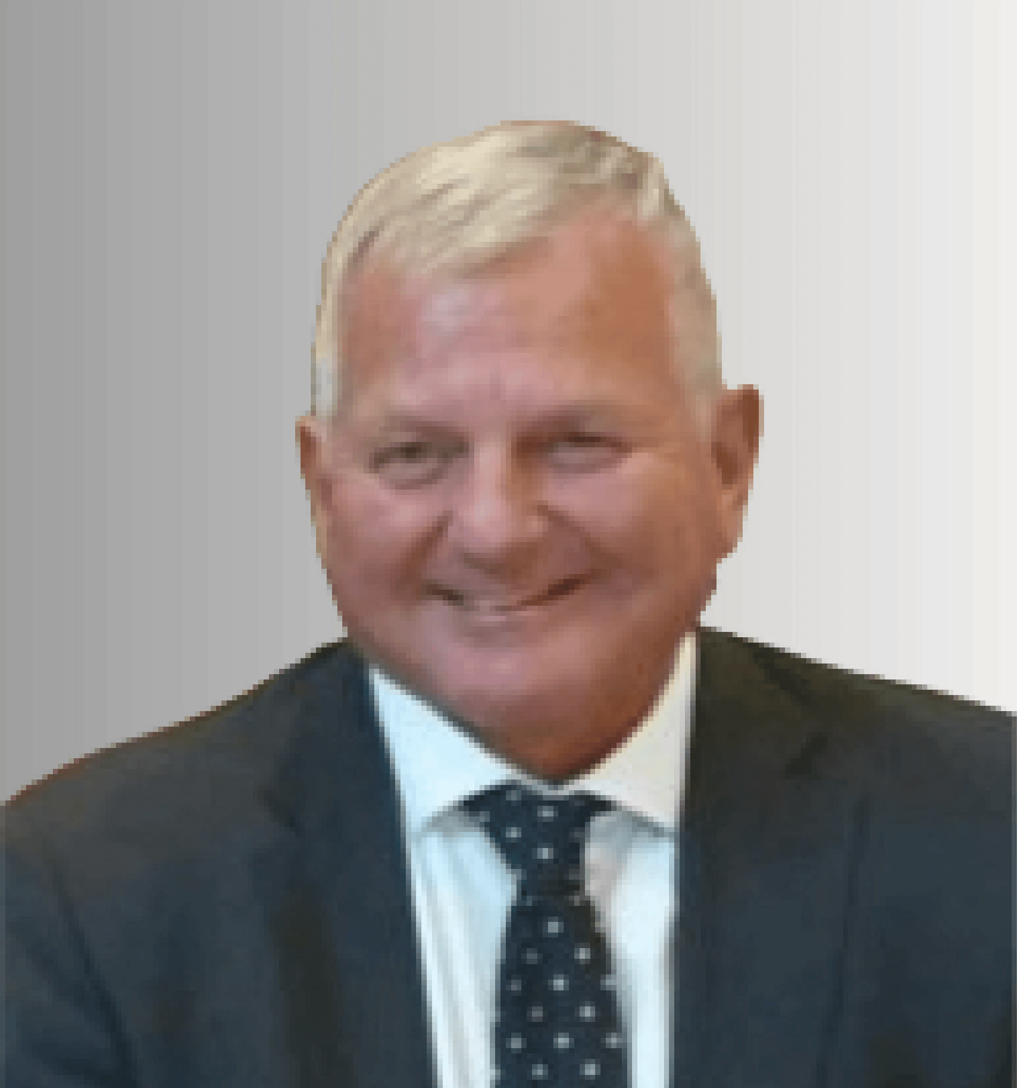A smiling man with curly hair is sitting at a desk, looking at the camera, with a laptop and a lamp in the background.