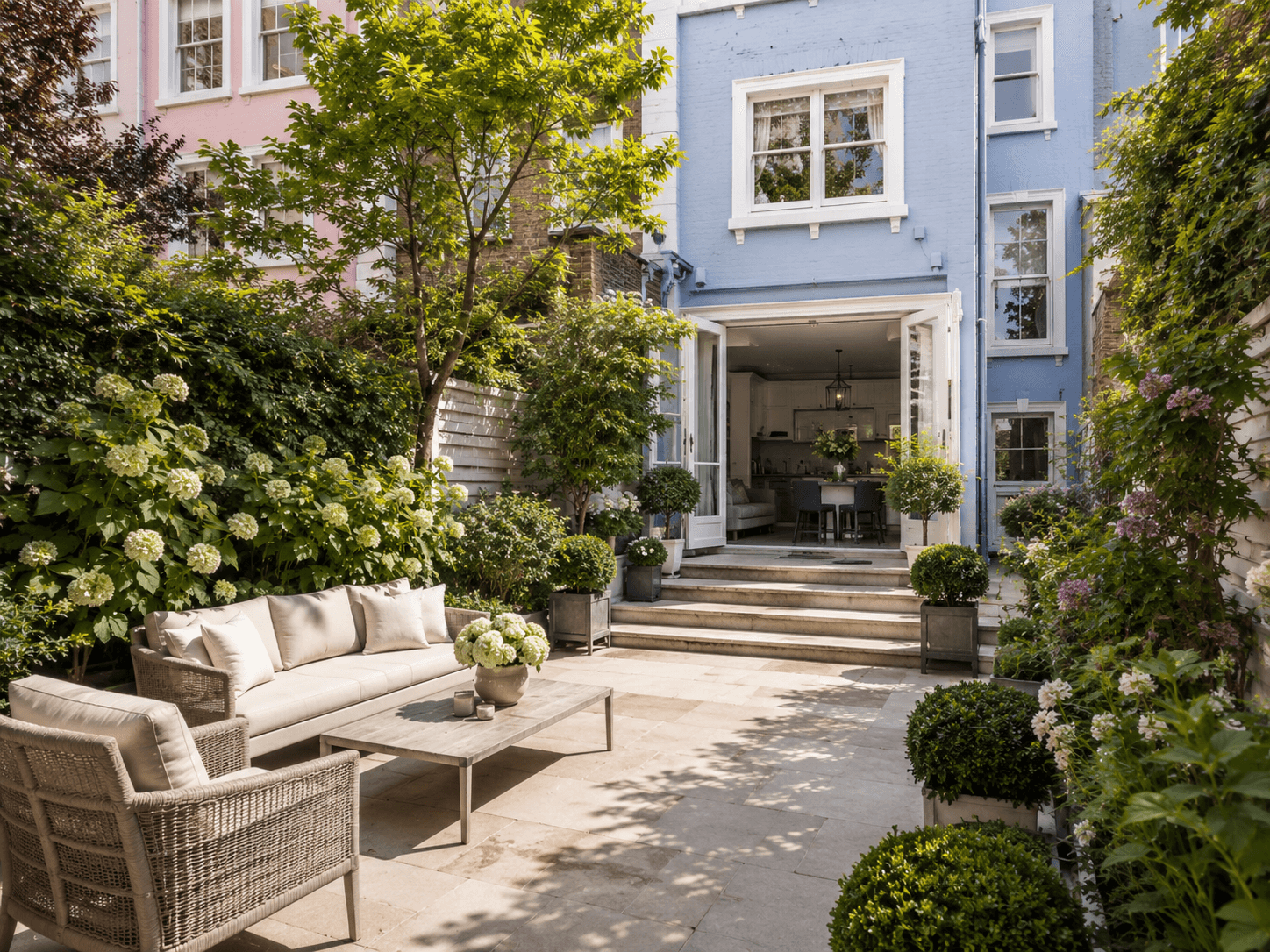 Private rear garden terrace behind a pastel blue Notting Hill townhouse, featuring stone paving, rattan outdoor seating, hydrangeas, lush greenery, and open doors to the kitchen.
