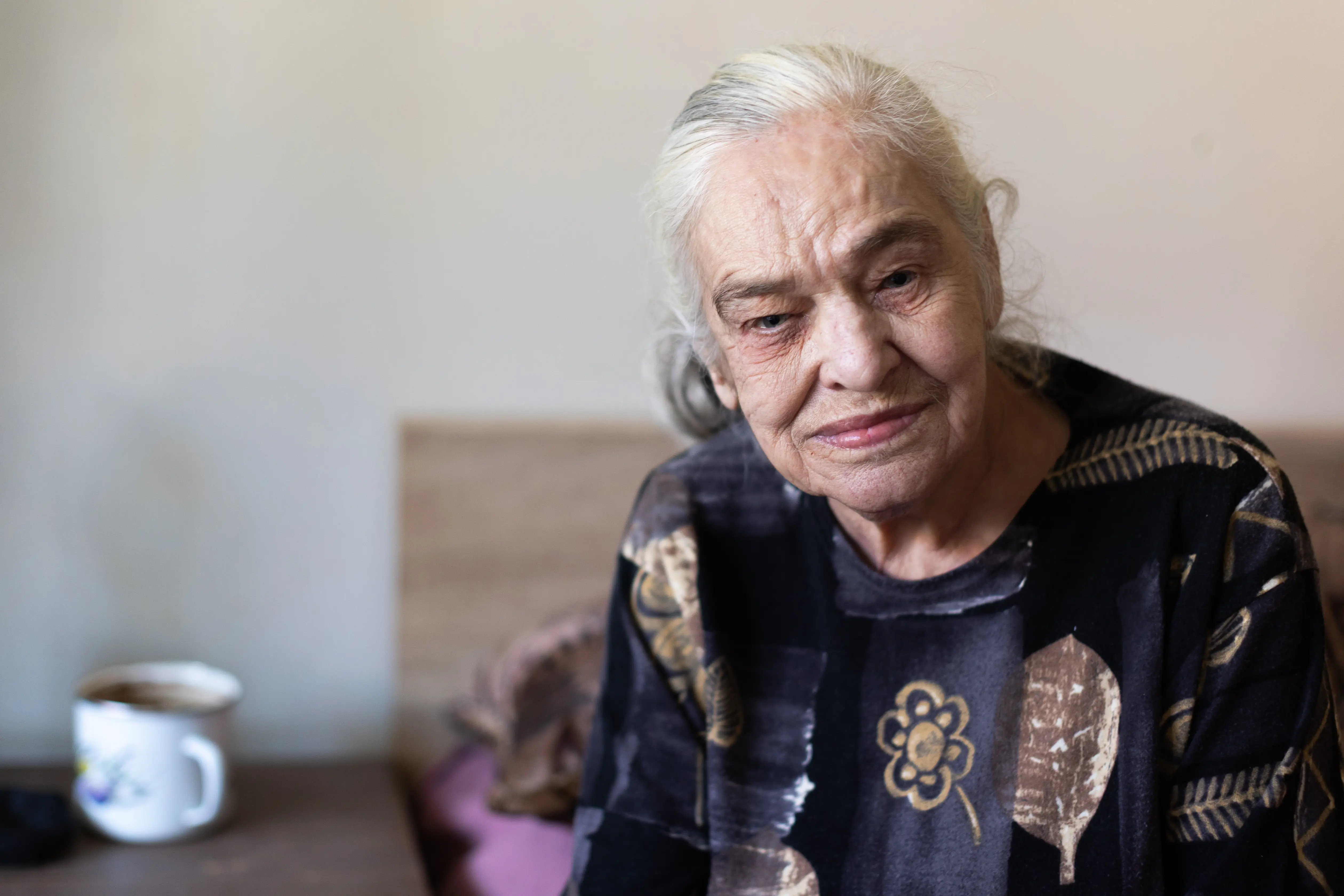 Starsza,siwa, uśmiechnięta pani w ciemnej bluzce siedzi na skraju łóżka, patrząc w obiektyw.
