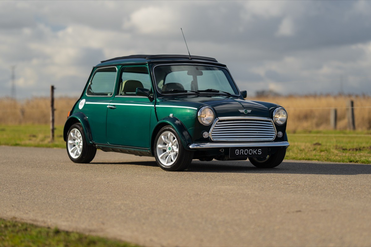 A vintage green car parked on a winding road with fields and cloudy skies in the background.
