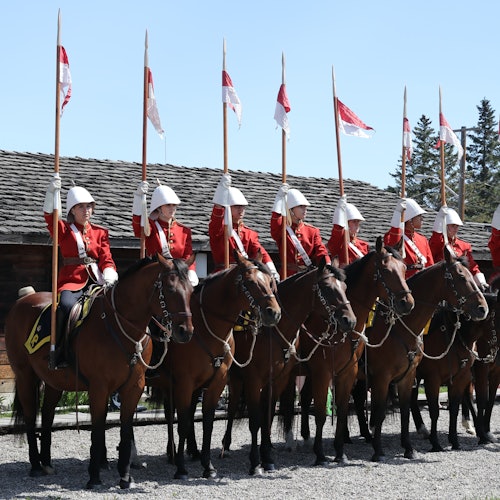 A line of uniformed horseback riders in red jackets and white helmets hold flags with red and white pennants outside a wooden building.